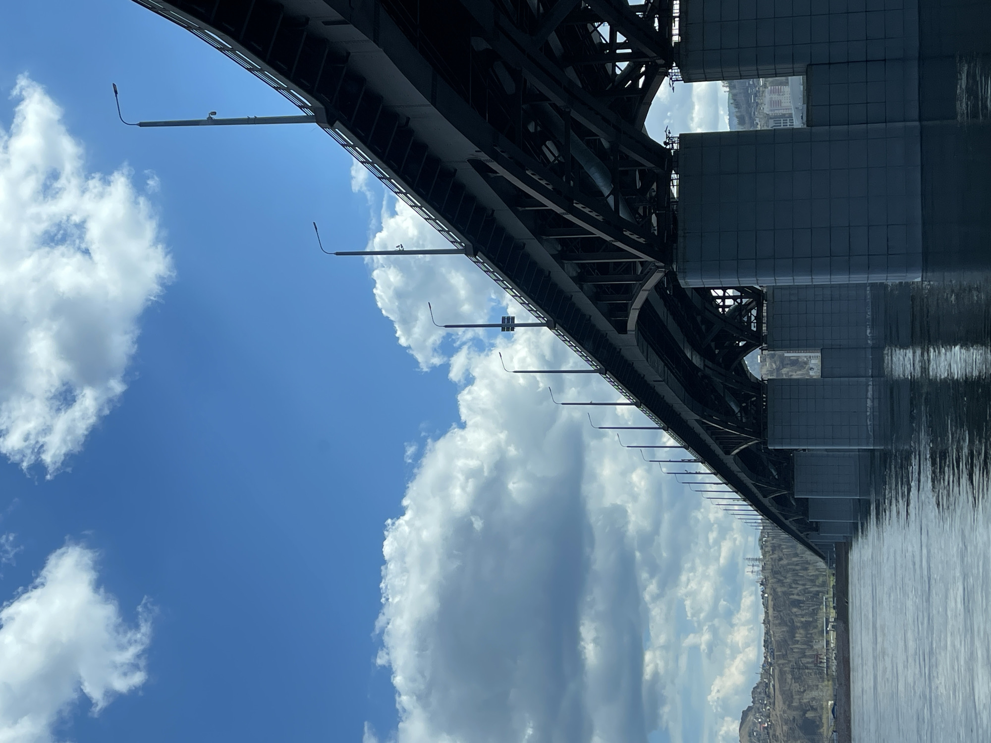 The image depicts a low-angle view under a bridge, revealing a sky with white clouds and the bridge's structure. The location appears to be Krasnoyarsk, Russia, due to the visible bridge architecture and surrounding landscape. The bridge's underside is metal and includes supporting pillars. Reflections in the water below indicate a river or lake. Small light fixtures are mounted along the bridge structure. A distant cityscape is visible through the bridge's openings. The image is oriented sideways.