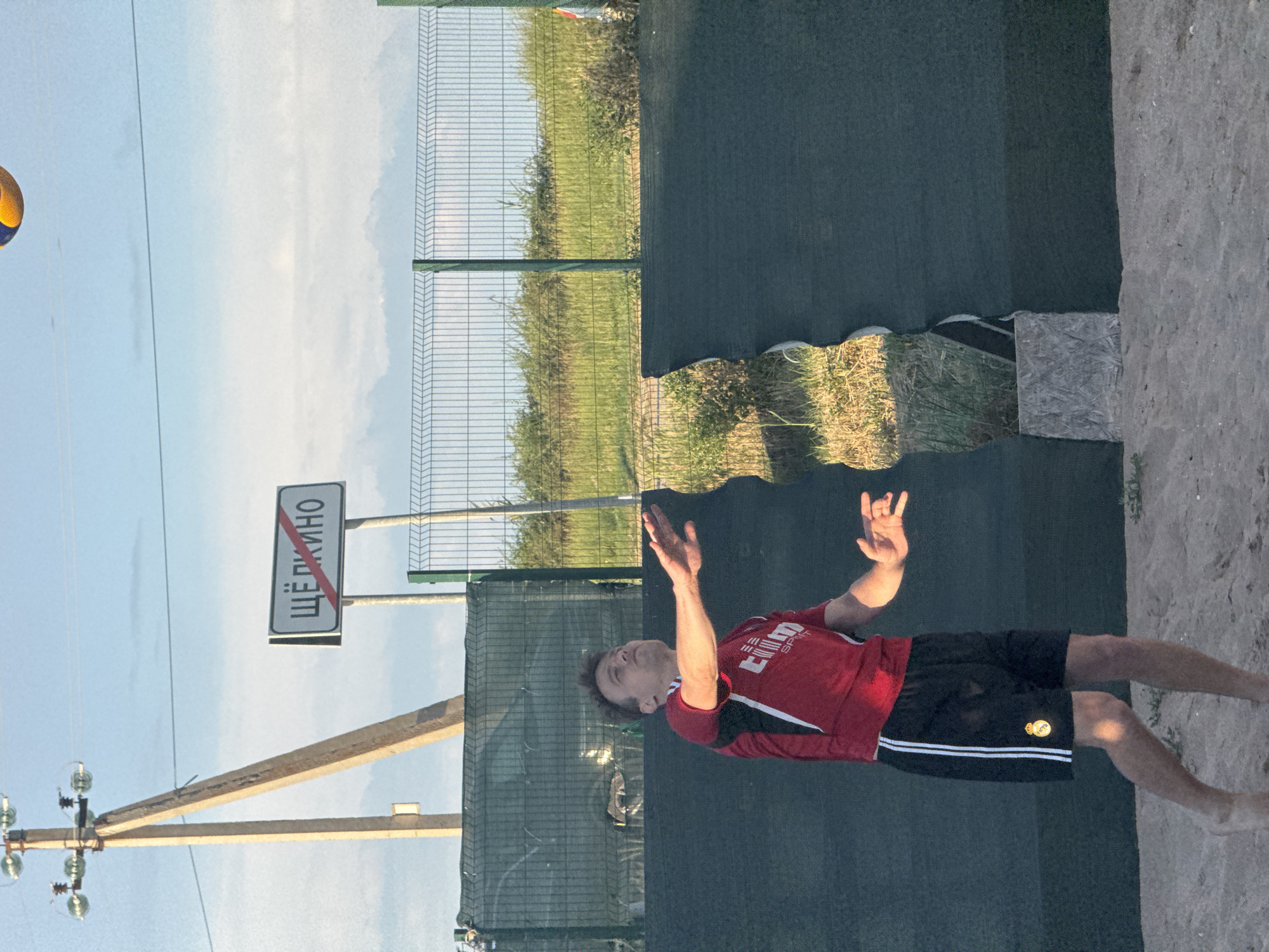 In the city of Shchelkino, Ukraine, a young man in athletic attire is playing volleyball on a sandy court. He is mid-action, with his arms outstretched, about to hit or having just hit a volleyball that is visible in the upper portion of the image. The man wears a red and black jersey with white trim and black shorts with white stripes and a gold emblem. The court is bordered by a dark green mesh fence, and a metal fence is visible further in the background. Beyond the fences, grassy fields and bushes are seen under a bright sky with scattered clouds. A wooden utility pole with insulators and a sign with Russian text, "ЩЁЛКИНО" (Shchelkino) and a red diagonal line, indicating a prohibition or end of the town, stands to the left of the man, near the edge of the court. The overall scene captures a moment of outdoor recreational activity in what appears to be a suburban or rural setting within Shchelkino.