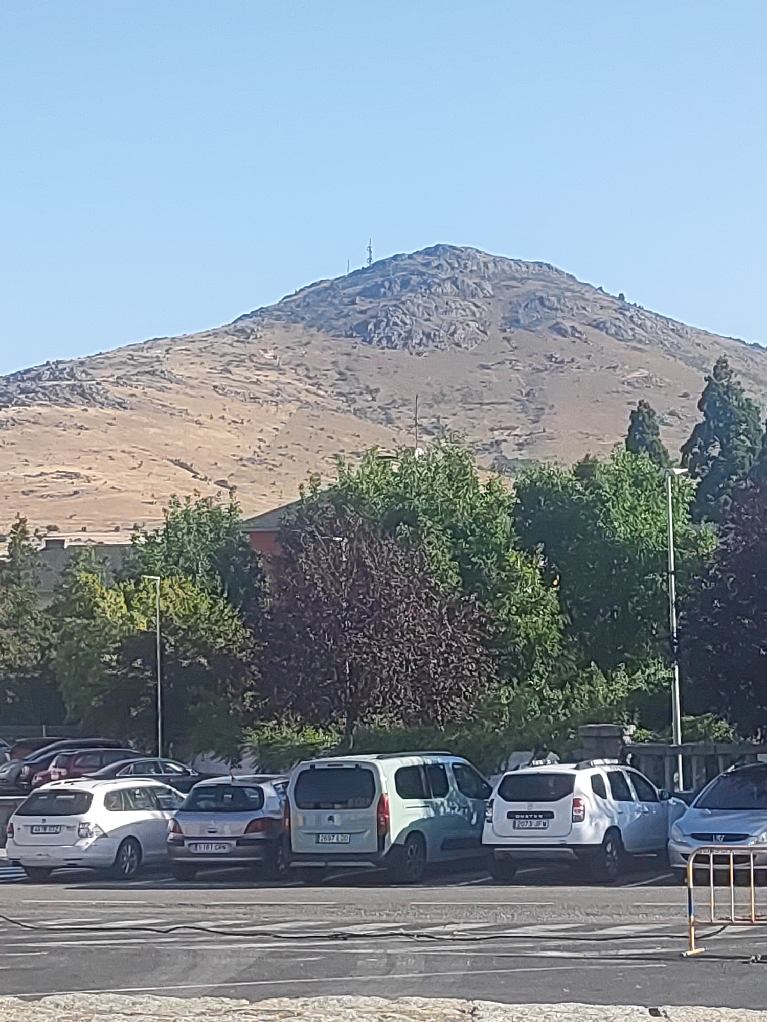 The image shows a mountain range in the background under a clear blue sky. The mountain slopes are covered in dry, tan vegetation with rocky outcrops and scattered trees. A few antennas are visible at the summit of the mountain. In the middle ground, there is a dense line of trees with green and some dark red foliage. Behind the trees, parts of buildings are visible, including a red-tiled roof. In the foreground, several cars are parked in a lot. These include a white station wagon, a silver hatchback, a white van, and a white SUV. The parking lot is paved, with some visible road markings. The scene appears to be a scenic overlook or a parking area near a natural landscape. The location is identified as being in the city of San Ildefonso, Spain.
