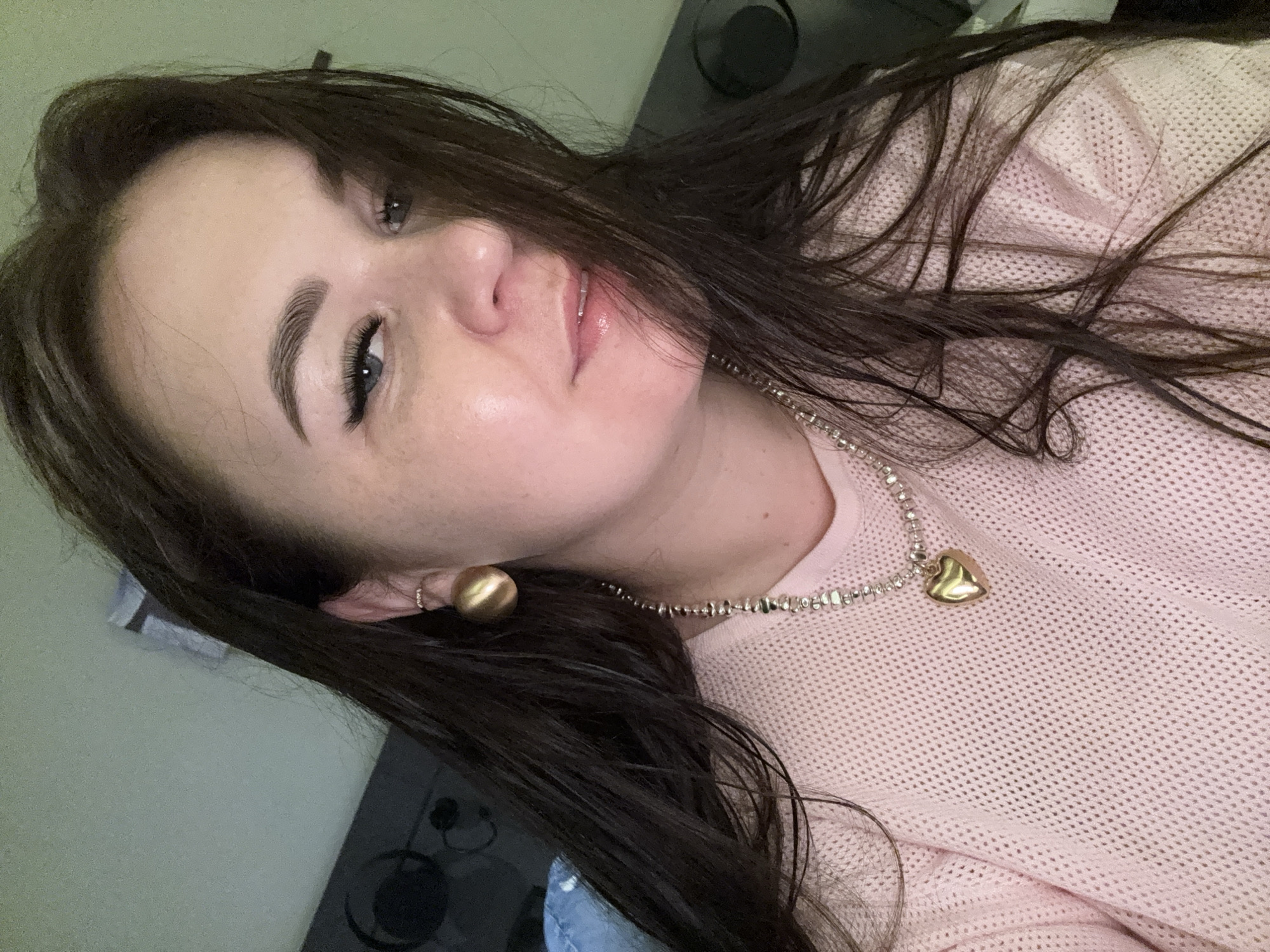 The image is a close-up, selfie-style photograph of a woman with long, dark brown hair. She is wearing a light pink, textured sweater, a beaded necklace with a gold heart-shaped pendant, and a large, round gold earring in her visible ear. Her makeup includes defined eyebrows, winged eyeliner, and lip gloss. The background appears to be an indoor setting with a green wall and dark furniture. There is no discernible location in Alanya, Turkey, within the image.