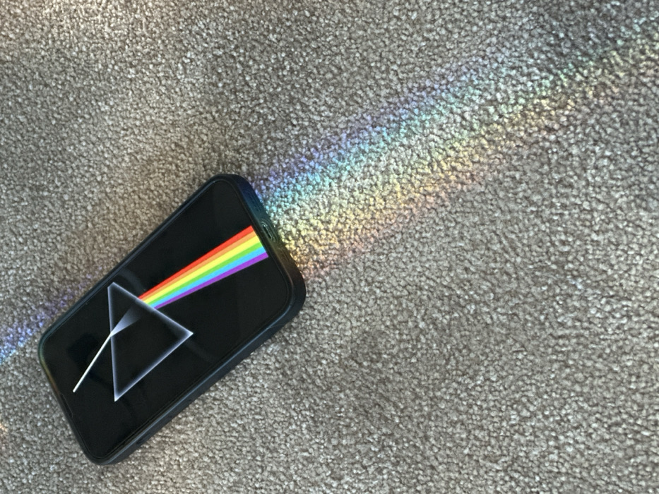 A black smartphone, positioned diagonally, rests on a light-colored, tightly looped carpet. The phone's screen is illuminated, displaying the "Dark Side of the Moon" album artwork: a white triangular prism refracting a white light beam into a rainbow spectrum that exits towards the bottom right of the screen. Coinciding with the on-screen display, a vivid, real-world spectrum of light, featuring red, orange, yellow, green, blue, indigo, and violet hues, emanates from the bottom right edge of the physical smartphone and projects diagonally across the carpet towards the lower right corner of the frame. The carpet surface is consistent throughout the visible area. No individuals or additional objects are present. The precise location within the city of Paris, France, cannot be determined from the visual information provided.