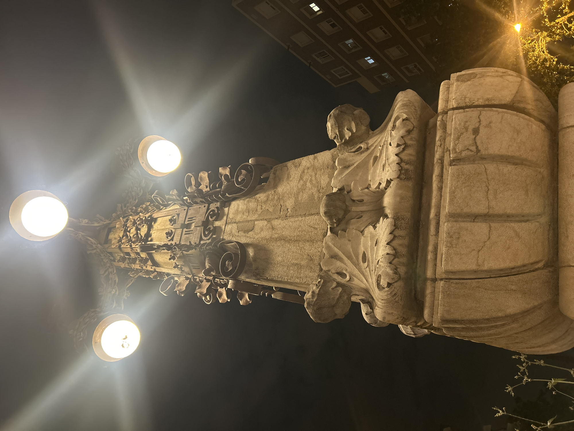 A low-angle, nighttime photograph depicts a street lamp in Barcelona, Spain. The lamp has three bulbs that emit bright white light. The pole is made of ornamented concrete. The lamp post base is detailed with baroque adornments. The night sky appears black. An apartment building is visible in the background.
