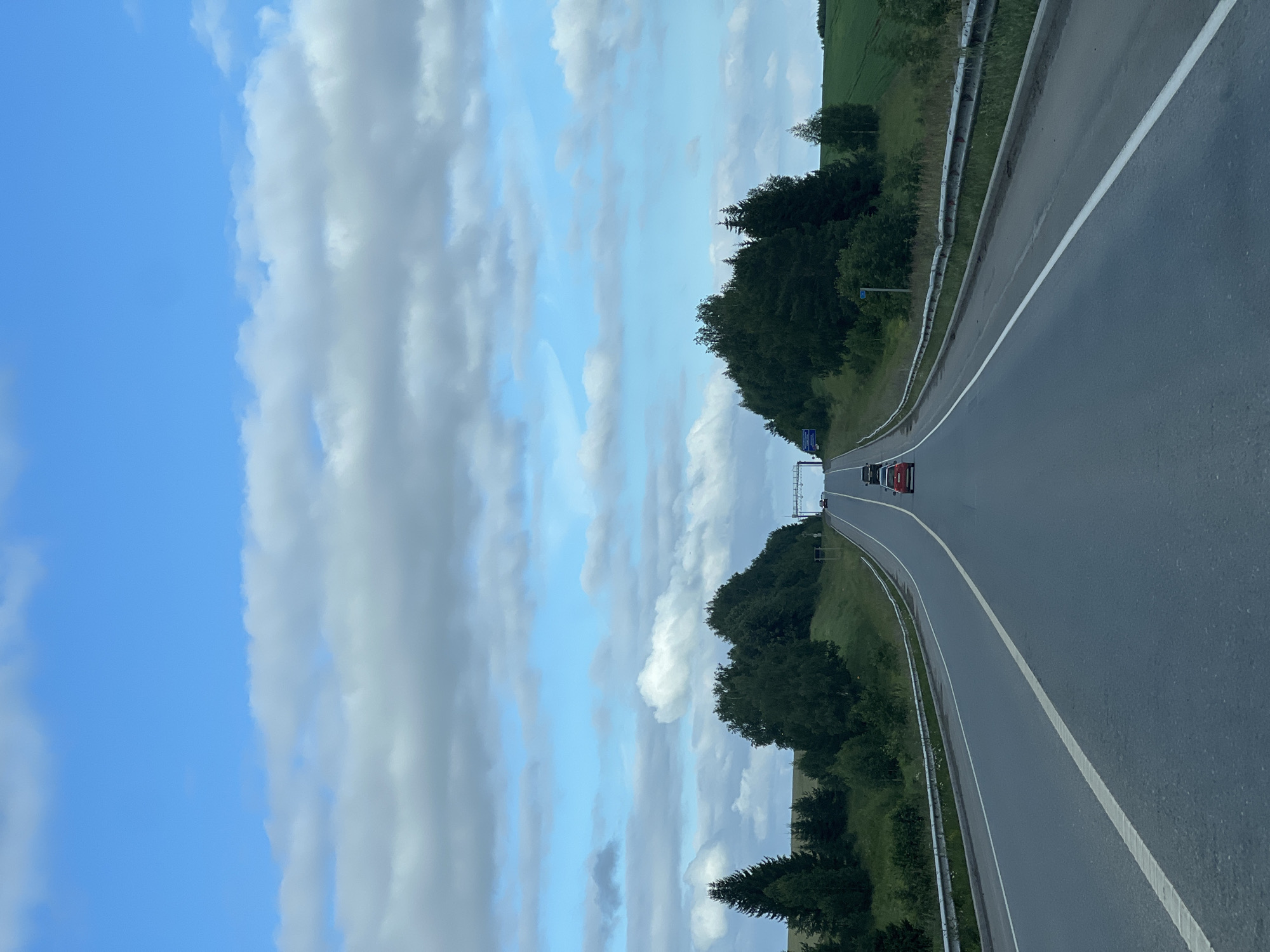 This image depicts a rural highway landscape, likely located near Verkhne-Pozim', Russia, under a partly cloudy sky. The perspective is from a moving vehicle, looking forward along a two-lane asphalt road that curves gently to the right as it ascends a slight incline.
The road is marked with a solid white line on the right edge and a broken white line separating the lanes. Guardrails, constructed of metal, run along both sides of the road. On the left side of the road, a dense line of dark green trees, including coniferous and deciduous varieties, borders a grassy embankment. On the right side, the terrain slopes upwards, with more trees and patches of green fields visible.
Several vehicles are present on the road. In the foreground, a red car is visible in the right lane. Further ahead, in the distance, more cars can be seen, including what appears to be a black SUV in the left lane. Overhead, a highway sign structure with what looks like electronic messaging capabilities is positioned above the road in the distance. The sky is a bright blue, interspersed with scattered, fluffy white and grey clouds. The overall impression is one of a typical summer day on a highway in a wooded, rural area.