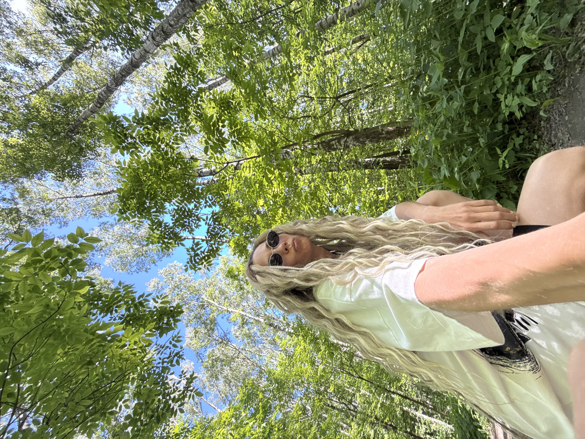 This image, captured in Rusanovo, Russia, depicts a low-angle view within a sunlit forest. A woman with long, light blonde, wavy hair and oval dark sunglasses occupies the lower left and central foreground, looking upwards and to the right with pursed lips. She wears a loose white t-shirt with a dark graphic design on her left side and is crouched or sitting on a dirt path. Her right arm is extended towards the camera, and her left hand rests near her thigh. The background features a dense canopy of vibrant green deciduous trees, including several tall trees with light-colored, possibly birch, bark, and darker-barked trees. Patches of clear blue sky are visible through the foliage. The undergrowth on the right side of the frame consists of various green plants, some with small yellow flowers. Bright daylight illuminates the scene.
