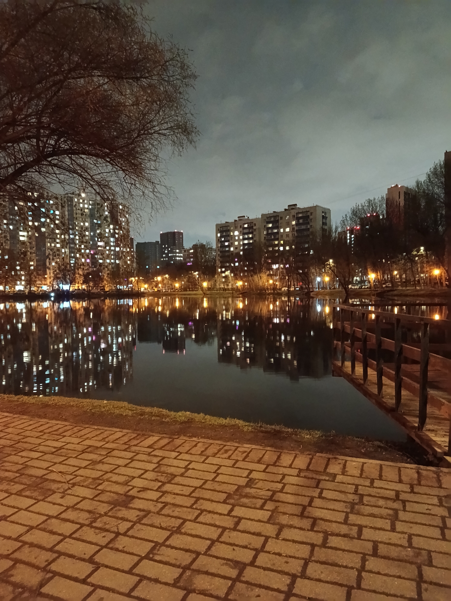 This image presents a nocturnal urban landscape in Moscow, Russia, dominated by the reflections of city lights on a body of water, likely a pond or lake.
In the foreground, a paved walkway of reddish-brown rectangular stones stretches across the bottom of the frame, with a thin strip of grass bordering the water's edge. To the right, a wooden railing or small bridge structure extends partly into the water. A large, bare-branched tree occupies the upper left quadrant, its dark branches silhouetted against the night sky.
Across the water, numerous multi-story apartment buildings are visible, their countless windows brightly illuminated, casting a warm, inviting glow. These lights, along with those from streetlights lining the far bank, create striking, elongated reflections that ripple across the water's surface, mirroring the urban architecture and vibrant activity within. The sky above is dark and overcast, providing a muted backdrop to the brightly lit cityscape. No people or specific activities are discernible, giving the scene a quiet, contemplative atmosphere. The overall impression is one of a serene, lived-in urban environment under the cover of night, with the warm artificial light contrasting with the natural darkness. No visible text is present.