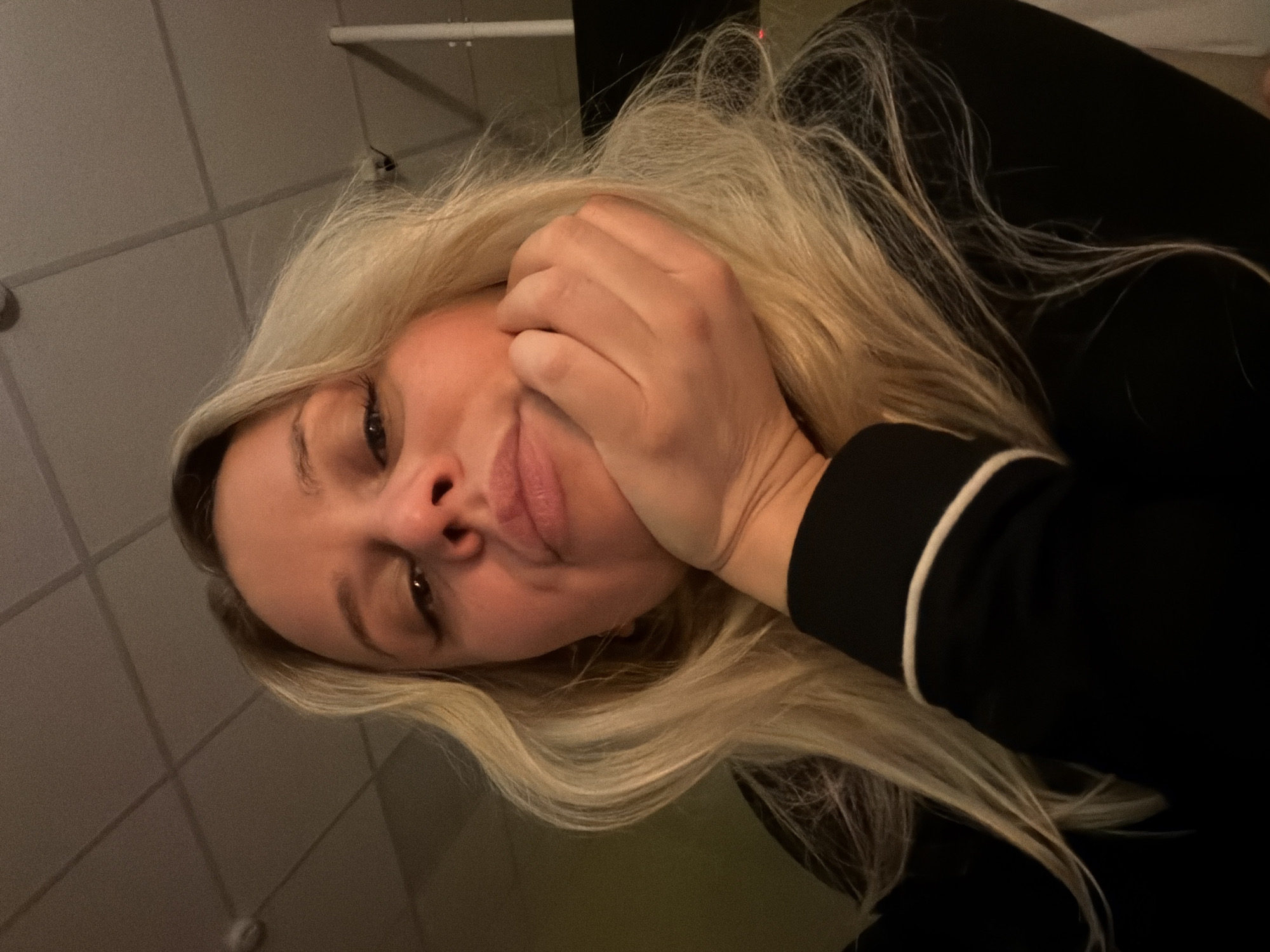 A close-up image depicts a female subject in an indoor setting located in Sarapul, Russia. The subject is Caucasian, with long, blonde hair falling over her shoulders. Her head is supported by her right hand, with fingers curled under her chin. She looks slightly upward towards the camera, her eyes defined with dark eyeliner and her lips exhibiting a subtle pout. She is wearing a dark, long-sleeved garment, with a white stripe visible on the cuff of her right sleeve. The background is dimly lit, revealing a light-colored suspended ceiling with grid lines. A round, light-colored object, consistent with a smoke detector or small light fixture, is visible on the ceiling in the upper left quadrant. The scene is illuminated by a warm, low-level light source.