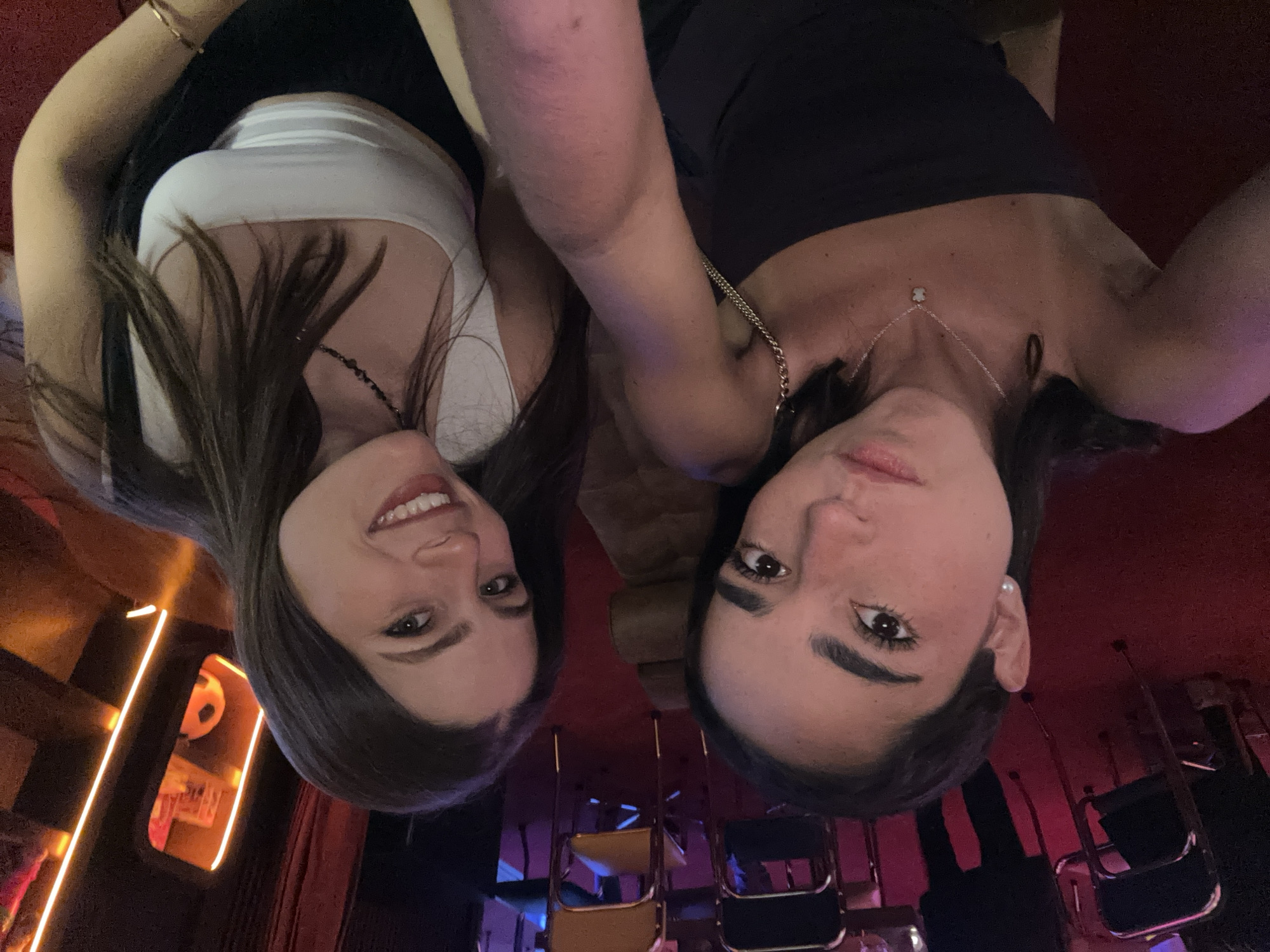 This vertical, inverted image captures two young women, likely a selfie taken from above, with their faces positioned at the bottom of the frame and looking upwards. The woman on the left has long dark hair, wears a white sleeveless top, a dark beaded necklace, and a gold bracelet on her left wrist; she is smiling broadly. The woman on the right has dark hair, wears a dark strapless top, a silver chain necklace with a small bear-shaped pendant, and a pearl earring on her right ear, with a more subtle smile or neutral expression.
The setting appears to be an indoor space, possibly a dimly lit bar, restaurant, or lounge, characterized by atmospheric lighting with dominant red and purple hues, and some warm orange light accents. In the background, chairs with metal frames, some black and some yellow/brown, are visible. On the left side of the frame, there are linear orange lights and a spherical object resembling a ball, hanging within a window-like alcove. The activity is posing for a photo. There is no visible text.