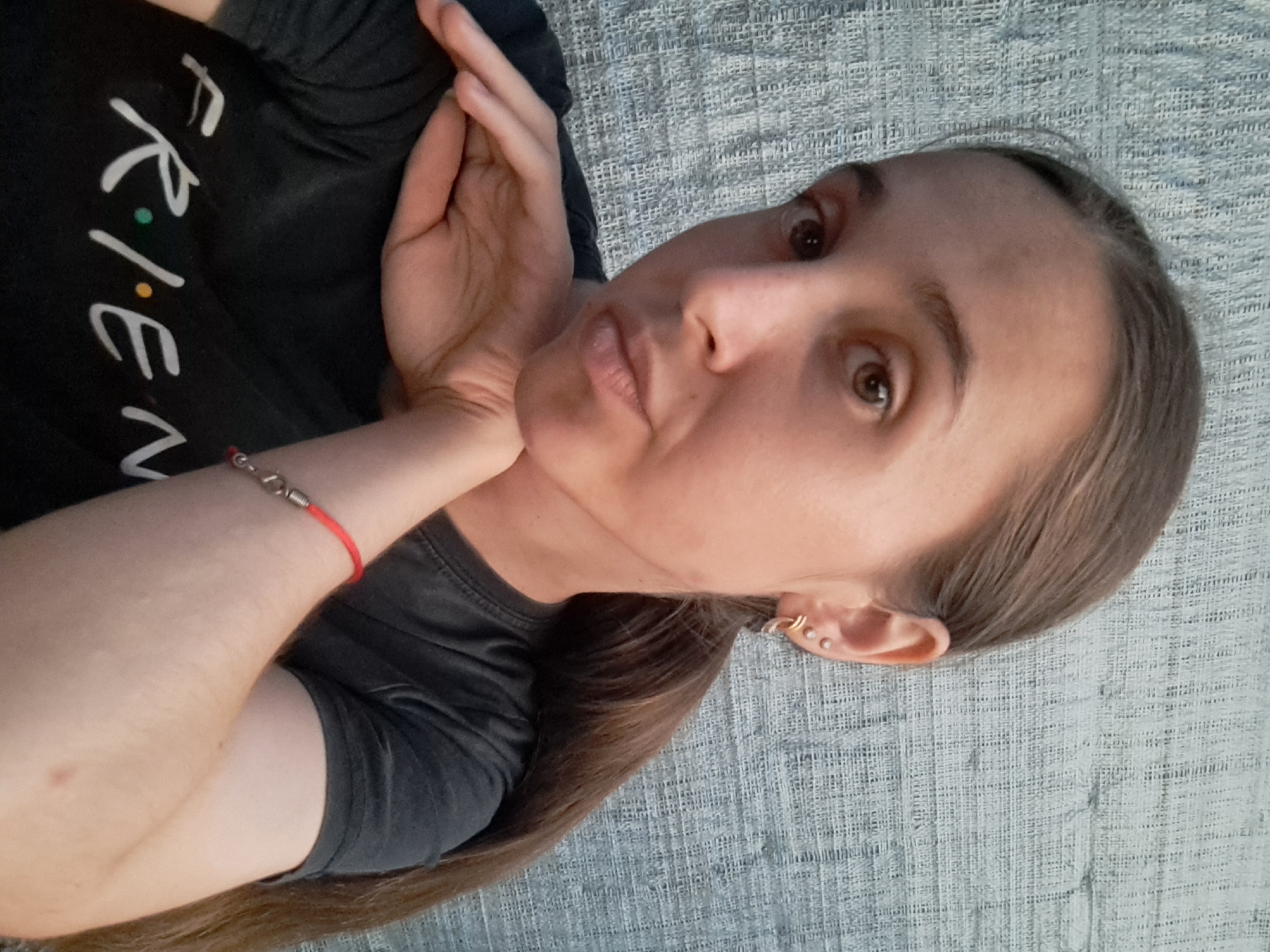 A young woman is captured in a close-up, selfie-style photograph. She has light brown hair pulled back into a ponytail and is looking directly at the camera with a neutral expression. Her right hand is resting on her chin, and she is wearing a dark t-shirt with the word "FRIENDS" printed in white across the chest. She has several earrings in her left ear and a thin red bracelet on her right wrist. The background appears to be a textured, light blue surface. The image was taken in Vypasne, Ukraine.