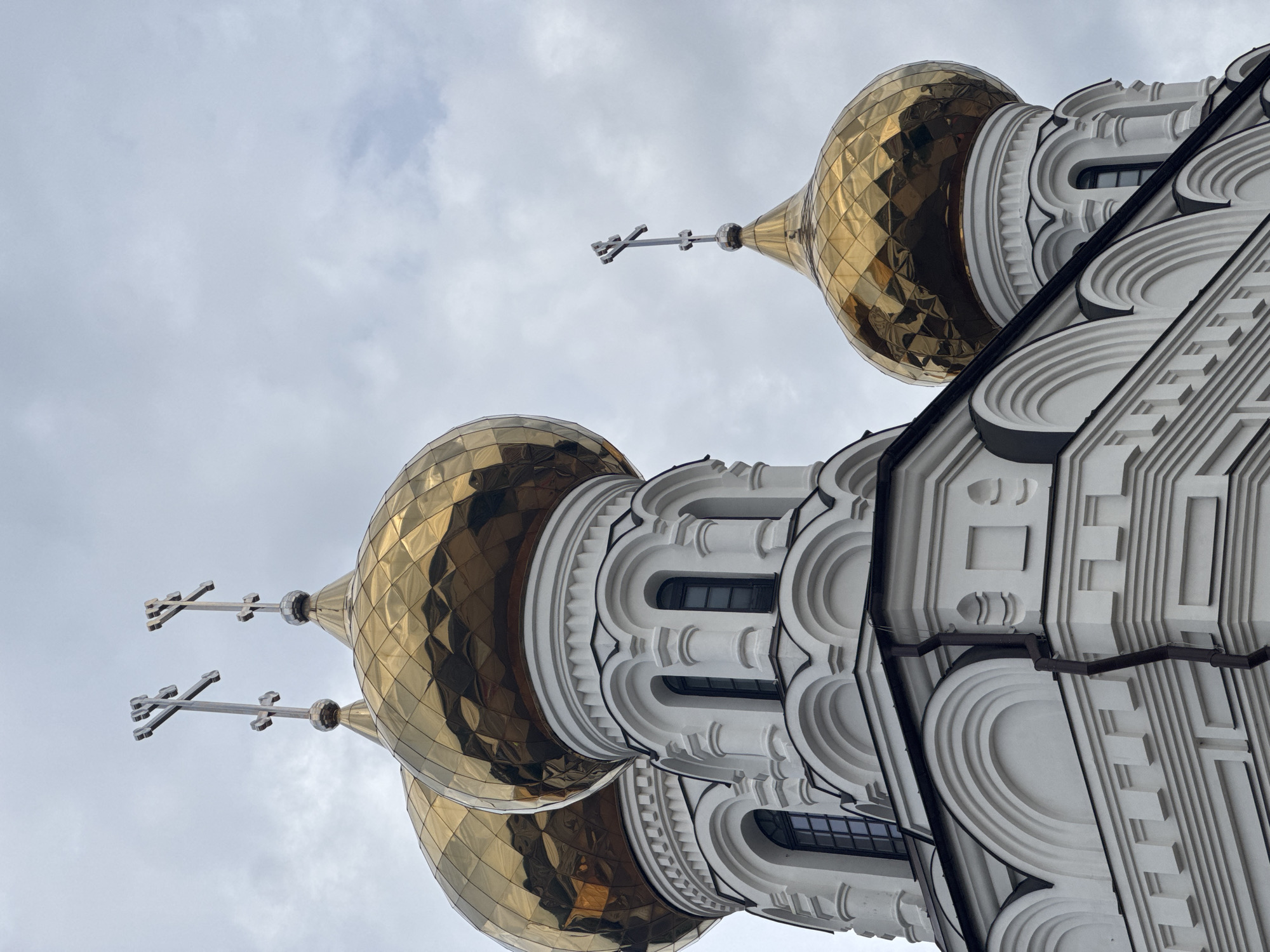 The image shows the golden domes of an Orthodox church against a cloudy sky. The architecture is white with arched windows and decorative detailing. Specifically, this is the Church of the Nativity of Christ, located in Tver, Russia. The photograph is a low-angle shot, emphasizing the grandeur of the church's domes and crosses. There are no people visible in the image. The overall mood is solemn and imposing, with the cloudy sky adding a sense of drama.