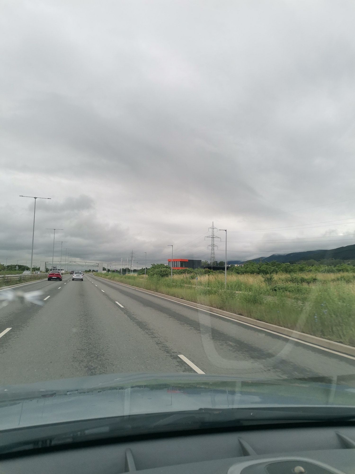 The image depicts a view from inside a vehicle traveling on a multi-lane highway under an overcast sky. The road ahead is wet, indicating recent rain. Several vehicles are visible on the highway, including a red car and a silver car in the left and center lanes, respectively. Streetlights line the left side of the road. On the right side, there is a grassy embankment with scattered green foliage. Further in the distance, a modern building with orange and dark gray sections is visible, along with power lines and transmission towers. Rolling hills or mountains are present in the background, shrouded in mist or clouds. The overall scene is one of a slightly dreary, overcast day on a highway.