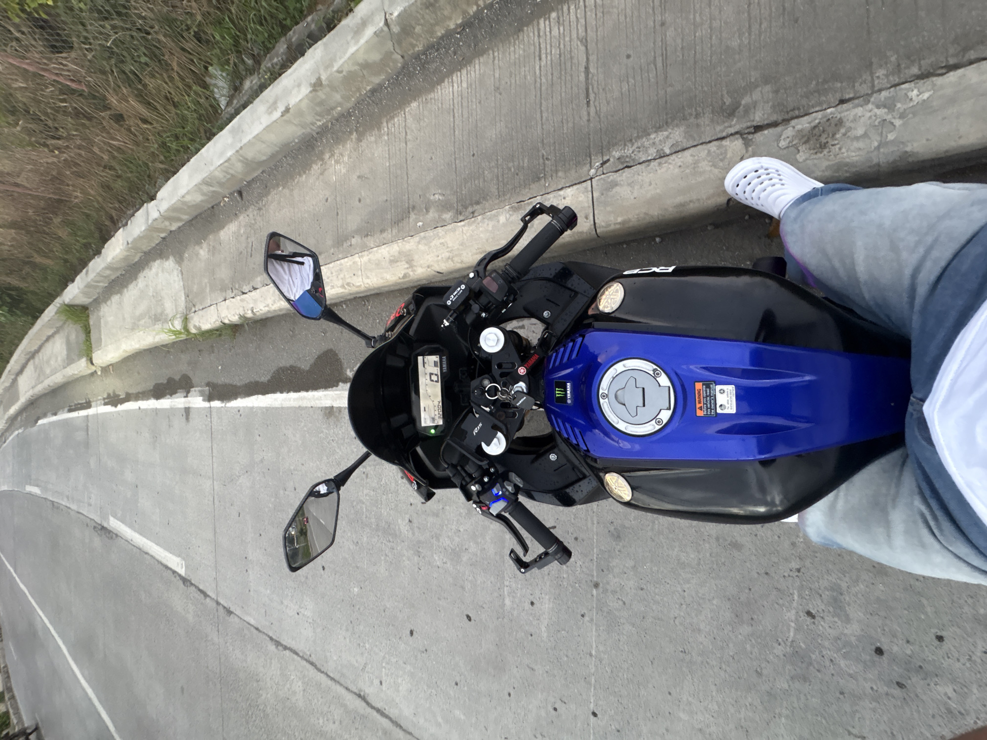 The image captures a first-person perspective of a person seated on a blue and black Yamaha R15 motorcycle, parked on the side of a road in Davao City, Philippines. The motorcycle is angled slightly towards the left, with its front wheel turned. The road features a white line dividing the lanes and a concrete curb runs along the right side, separating the road from overgrown vegetation. The sky appears overcast, suggesting it might be an early morning or late afternoon. The rider's legs, clad in blue jeans and white perforated slip-on shoes, are visible in the foreground. The motorcycle's digital dashboard displays "32004" kilometers. Reflections in the mirrors show a red vehicle in the distance and the rider's torso with a white shirt. The overall scene depicts a brief pause on a roadside journey.