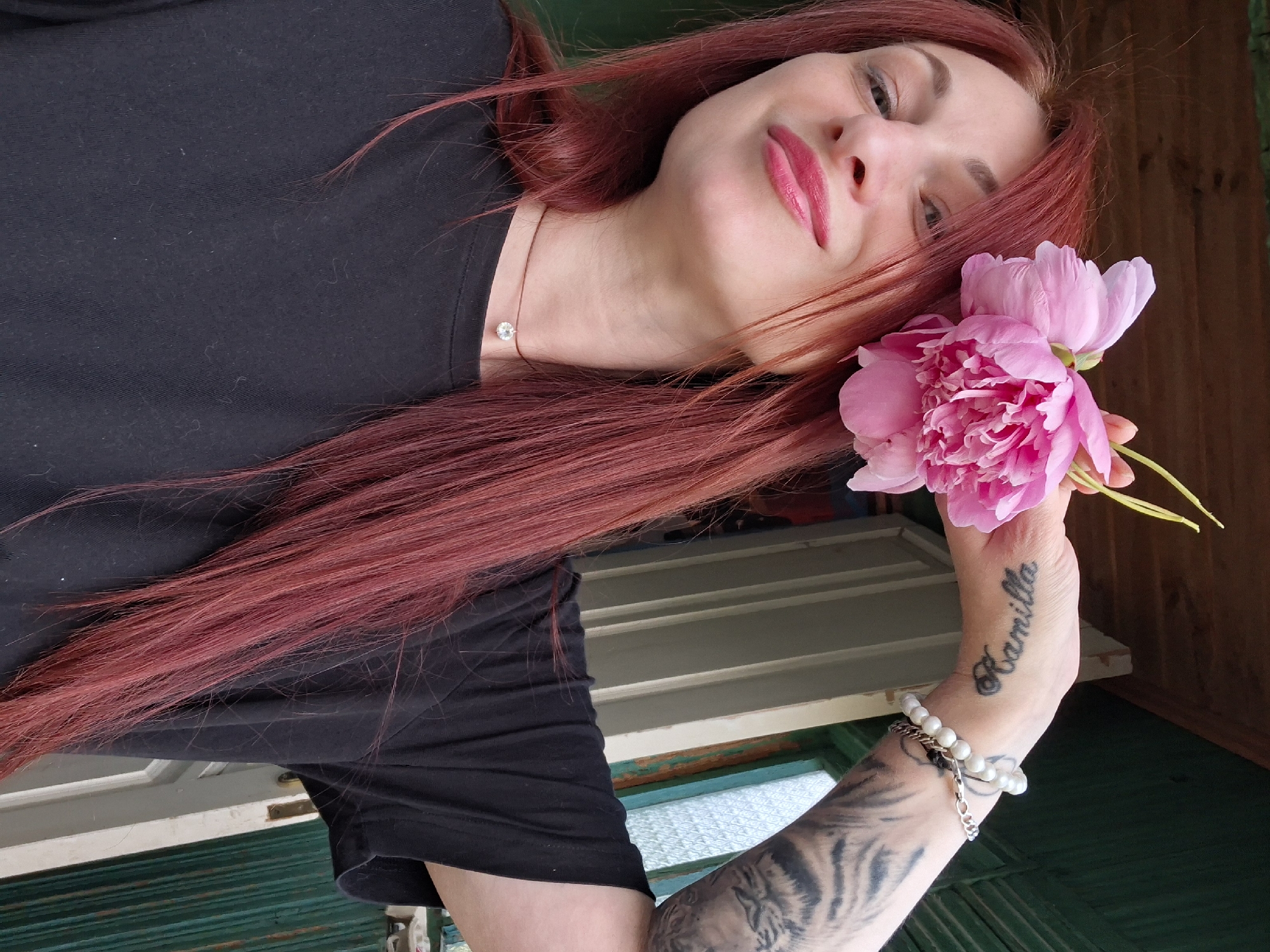 This is a portrait of a woman with long, reddish-brown hair and bright pink lipstick. She is wearing a black t-shirt and a delicate necklace with a small, clear pendant. She is holding a large, pink peony flower in her right hand, which is adorned with several bracelets and a tattoo on her forearm that spells out "Kamilla". The background suggests an outdoor setting, possibly a porch or an area with wooden walls and green corrugated metal panels. The lighting appears natural, indicating it is daytime, and the atmosphere is relaxed and intimate. The woman's gaze is directed towards the camera, with a slight smile and an element of playfulness.