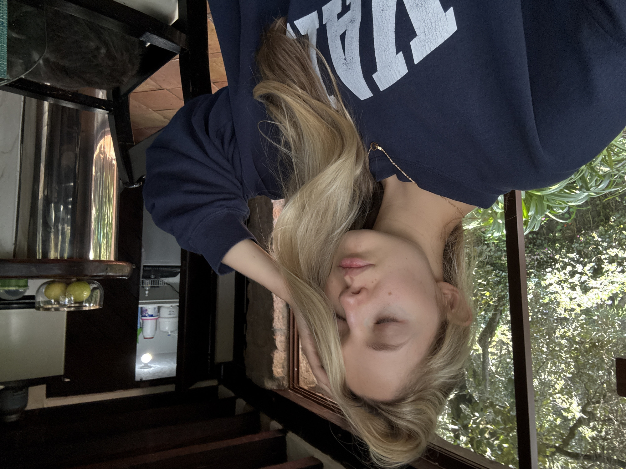 The image shows a young woman with blonde hair touching her head, with her eyes closed. She is wearing a dark blue sweatshirt with white lettering. She is indoors, in what appears to be a rustic kitchen or dining area, with exposed wooden beams and stone walls. Through a large open window or doorway, lush green foliage and trees are visible, suggesting an outdoor setting. In the background, there is a glimpse into a room with shelves stocked with various items, and a glimpse of a kitchen counter with a bowl of lemons. A dark-colored animal, possibly a cat, is visible in a basket in the lower right corner. The lighting suggests it is daytime. There is no discernible connection to the city of Ciudad de México, Mexico.