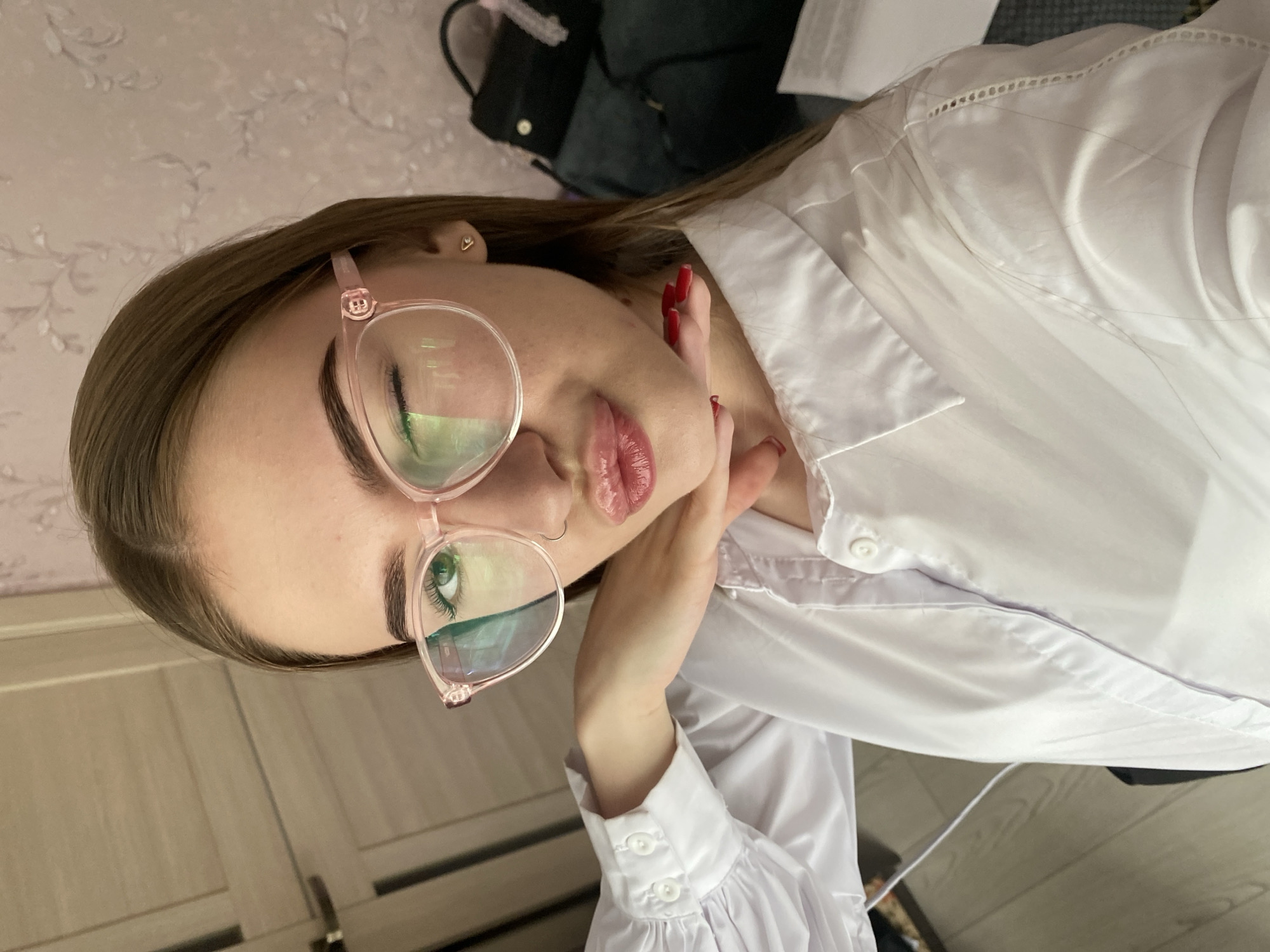 The image shows a young woman, identified as being in Dyakovo, Russia, taking a selfie. She is wearing a white collared shirt with ruffled cuffs and large, pink-rimmed glasses. Her left eye is closed in a wink, and her lips are pursed. She has a small nose ring and a heart-shaped earring. Her right hand is raised, with red-painted nails, resting on her chin. In the background, a wooden door and a patterned wallpaper are visible, as well as a dark bag. The lighting suggests an indoor setting, possibly a home.