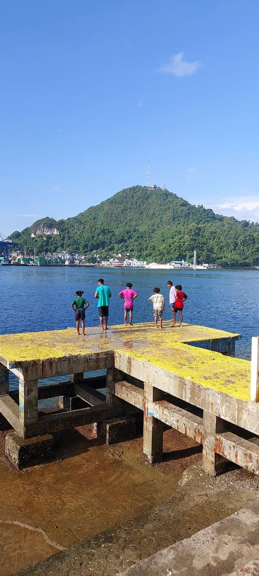 This is a vertical, eye-level photograph of a pier in Jayapura, Indonesia, with a mountain in the background. The pier is constructed of concrete with a bright yellow surface, possibly due to paint or debris. Five people, appearing to be children or teenagers, stand on the pier facing away from the camera, looking out at the water and the distant cityscape. One person wears a green t-shirt and dark shorts, another a teal t-shirt and shorts, a third a pink t-shirt and shorts, a fourth a striped t-shirt and shorts, and the last wears a red t-shirt and shorts. The water is a deep blue and calm, with several boats visible in the middle ground and a large green shipping crane on the left side of the frame. The mountain in the background is covered in lush green vegetation and features two communication towers near its summit, with the text "JAYAPURA CITY" visible between them. The sky is a clear, bright blue with a few wispy clouds. The foreground shows the concrete supports of the pier and the muddy water and ground beneath it, indicating a tidal area or port setting.