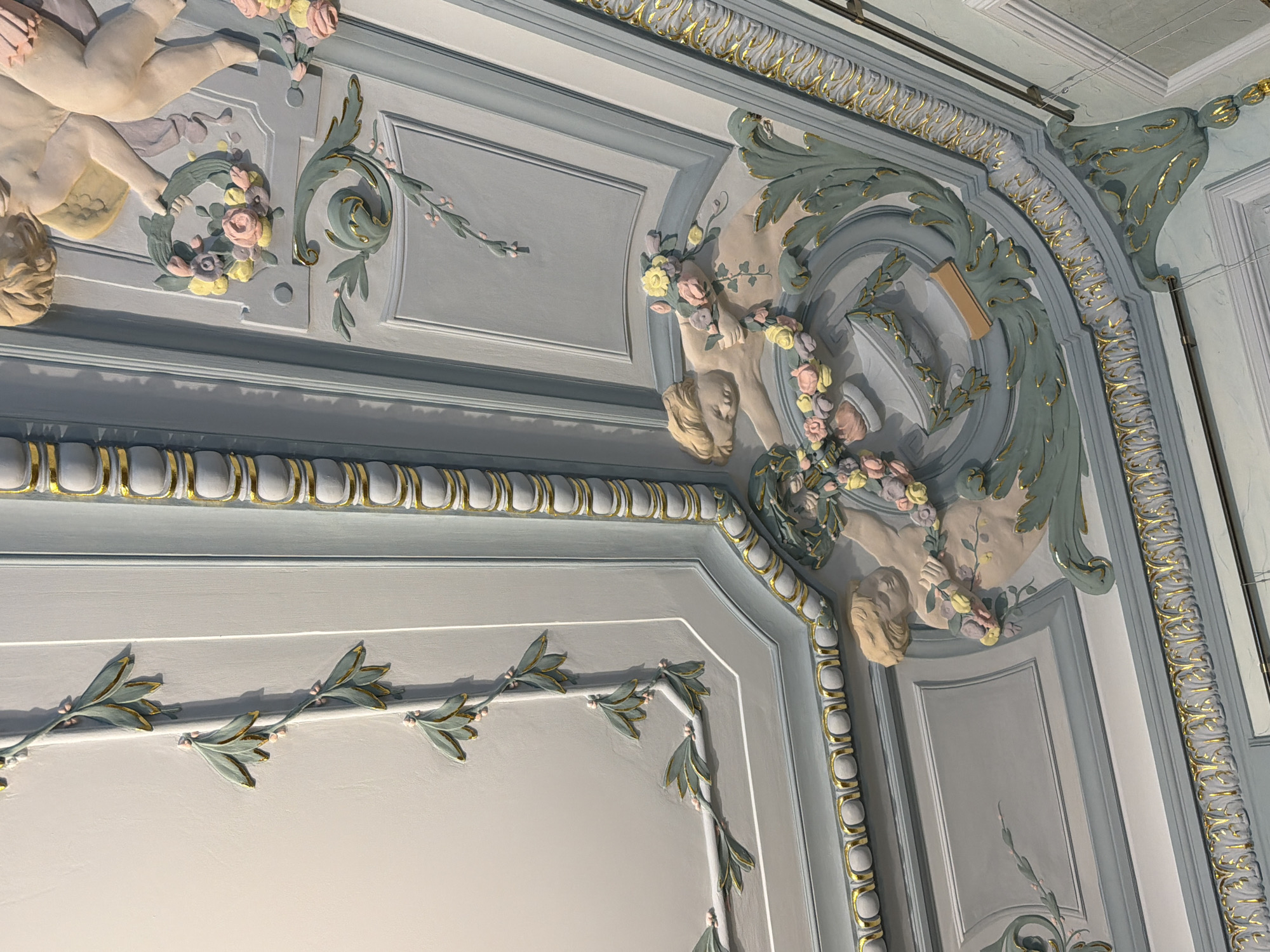 The image displays a detailed section of an ornate ceiling with plasterwork, likely within a historic building in Nizhny Novgorod, Russia. The ceiling is painted in soft pastel colors, predominantly light blue and cream, with accents of gold. The decorative elements include relief figures of cherubs or putti, draped in fabric and holding wreaths of pastel-colored flowers. Swirling acanthus leaves and floral motifs are also prominent, rendered in a pale green with gilded edges. The geometric structure of the ceiling, featuring recessed panels and raised moldings, is emphasized by the decorative plasterwork. The lighting suggests an indoor setting, possibly a grand hall or room.