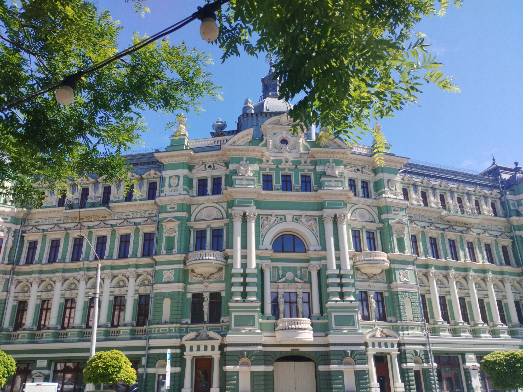 The image shows the facade of the London Hotel in Odesa, Ukraine. The building is a multi-story structure with a distinctive green and white color scheme and ornate architectural details, including columns, balconies, and sculptural elements. A dome-like structure tops the central portion of the building. Tree branches and street lighting fixtures partially frame the building in the foreground. The sky is clear and blue. The lower portion of the building includes entrances and ground-level details such as small, trimmed bushes and street signs.