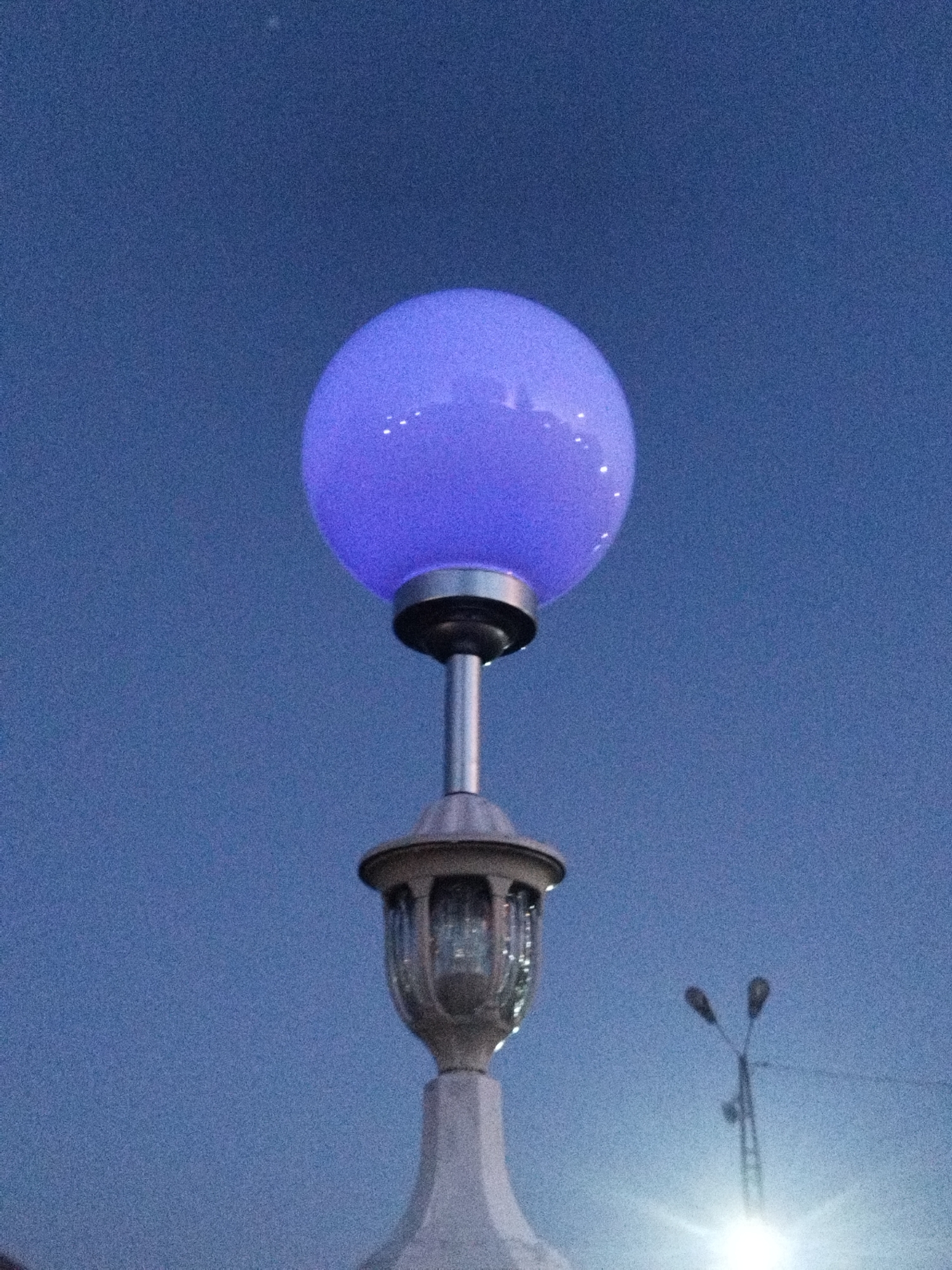 A purple globe lamp illuminates a clear blue sky. The lamp is mounted on a decorative lamp post that has a faceted glass lantern. In the distance, two streetlights stand tall against the sky. The image appears to be taken at dusk or dawn. There are no people or animals visible. The overall scene is one of urban illumination against a twilight sky.