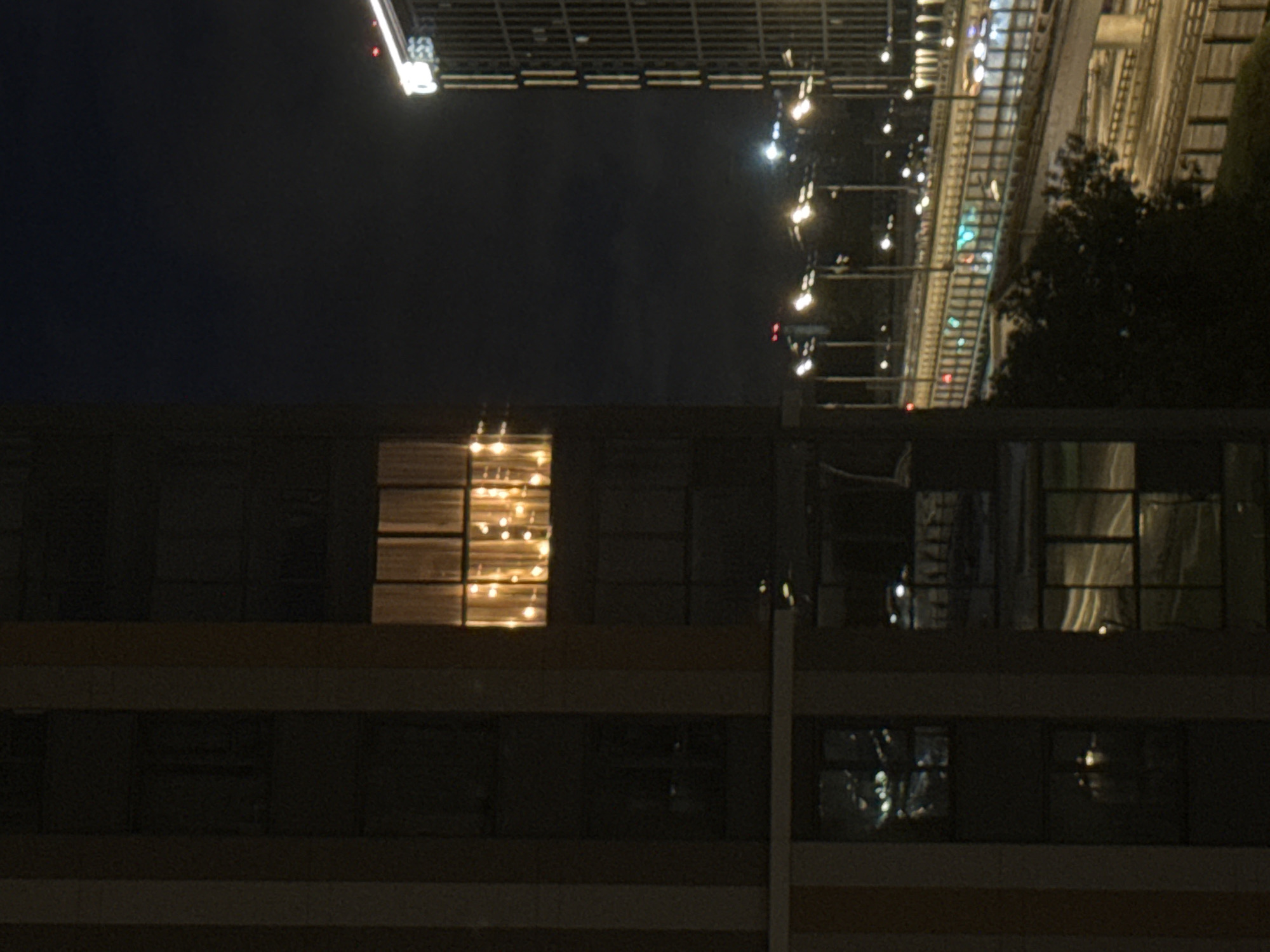 The image depicts a night scene in the city of Khimki, Russia, focusing on urban architecture and infrastructure. In the foreground, a portion of a tall building is visible, characterized by dark vertical elements and windows. One window on a higher floor is illuminated by a string of warm-toned lights, creating a festive or decorative effect. Below this, other windows reflect the night sky and distant city lights.
To the right of the foreground building, another modern high-rise structure stands, with its facade featuring vertical lines and illuminated windows. In the mid-ground, a multi-level highway or elevated road system is present, with cars visible as streaks of light, indicating movement. Streetlights line the roadway, casting bright pools of light. A dark, dense tree or cluster of trees occupies a section of the mid-ground, partially obscuring the view of the roadway. The background is dominated by the dark night sky, with faint lights from distant buildings and possibly clouds. The overall atmosphere is one of a nocturnal urban environment with a focus on light and shadow.