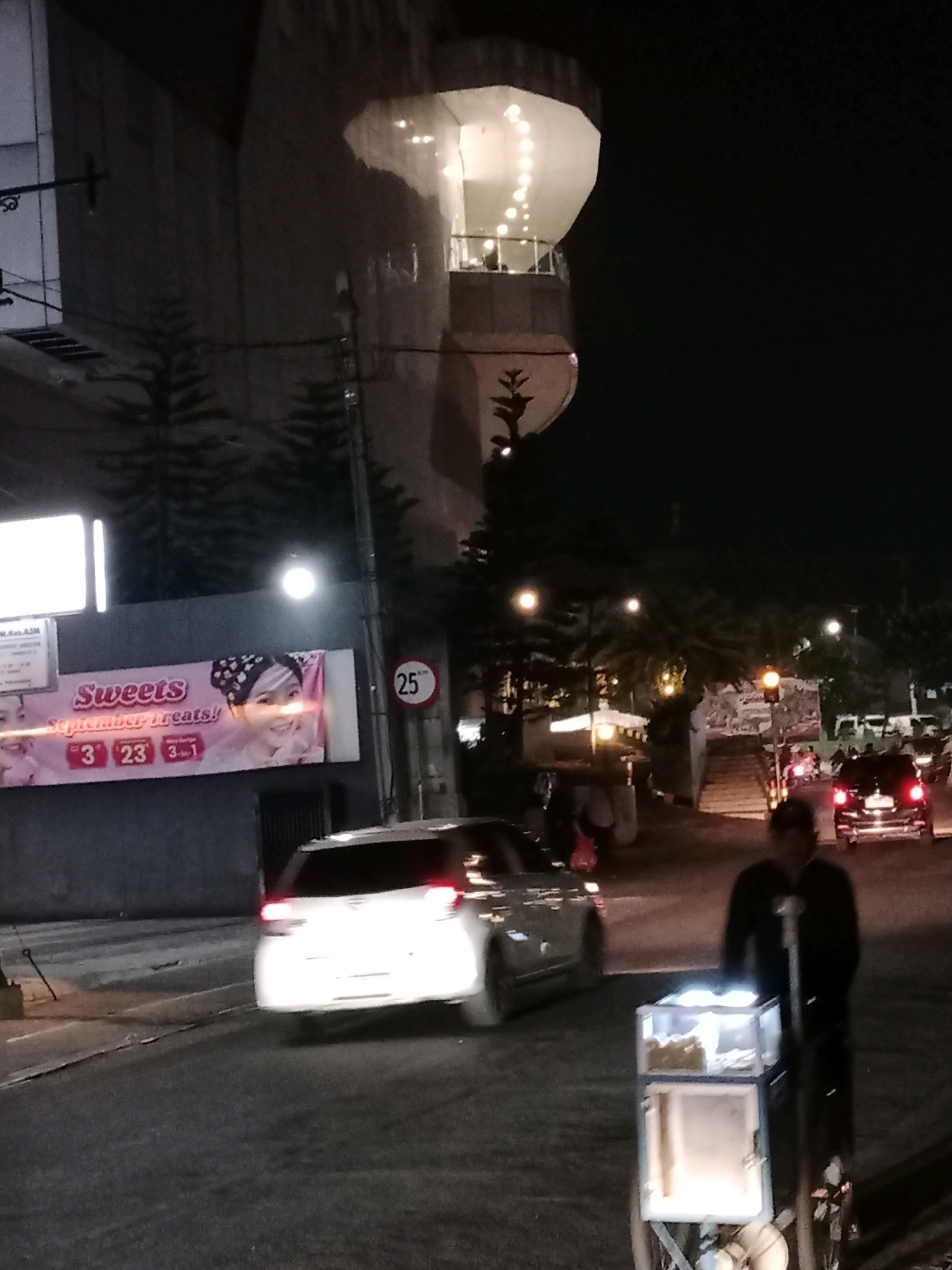 The image captures a night scene on a street in Cianjur, Indonesia, featuring a white hatchback car, a person pushing a food cart, and a billboard. The billboard, with the text "Sweets September treats!", displays images of women and dates "3", "23", and "3-1". A speed limit sign indicating "25" km/h is also visible. In the background, a multi-story building with illuminated sections and some trees are present. The street is paved, and the overall lighting suggests it's nighttime. The food cart is lit and appears to contain food items. The person pushing the cart is wearing dark clothing.