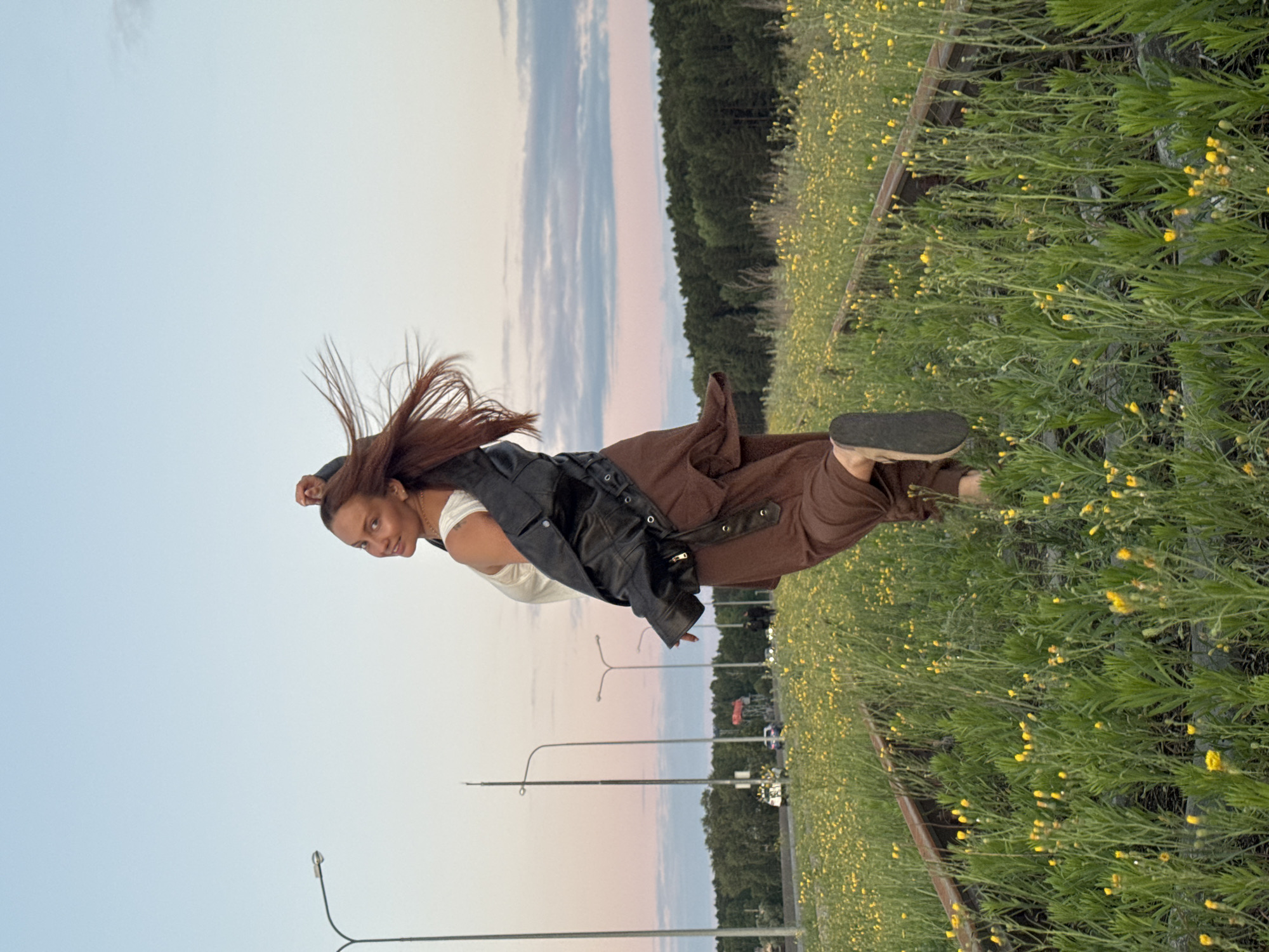 The photograph captures a young woman with long, reddish-brown hair, wearing a white tank top, a black leather jacket draped over her shoulders, and loose brown pants. She is mid-stride, seemingly running or jumping along a set of abandoned railway tracks that are overgrown with tall green grass and dotted with small yellow wildflowers. Her body is turned away from the camera, with her head looking back over her shoulder, a slight smile on her face. Her right arm is raised and bent at the elbow, and her right leg is extended backward. The setting appears to be on the outskirts of Chaikovsky, Russia, indicated by the presence of streetlights and a distant view of a road with some vehicles. Beyond the railway tracks, a dense line of dark green trees forms the horizon, silhouetted against a sky painted with soft hues of pink and blue, suggesting either dawn or dusk. The overall atmosphere is one of freedom and movement, set against a serene natural backdrop with urban elements visible in the distance.