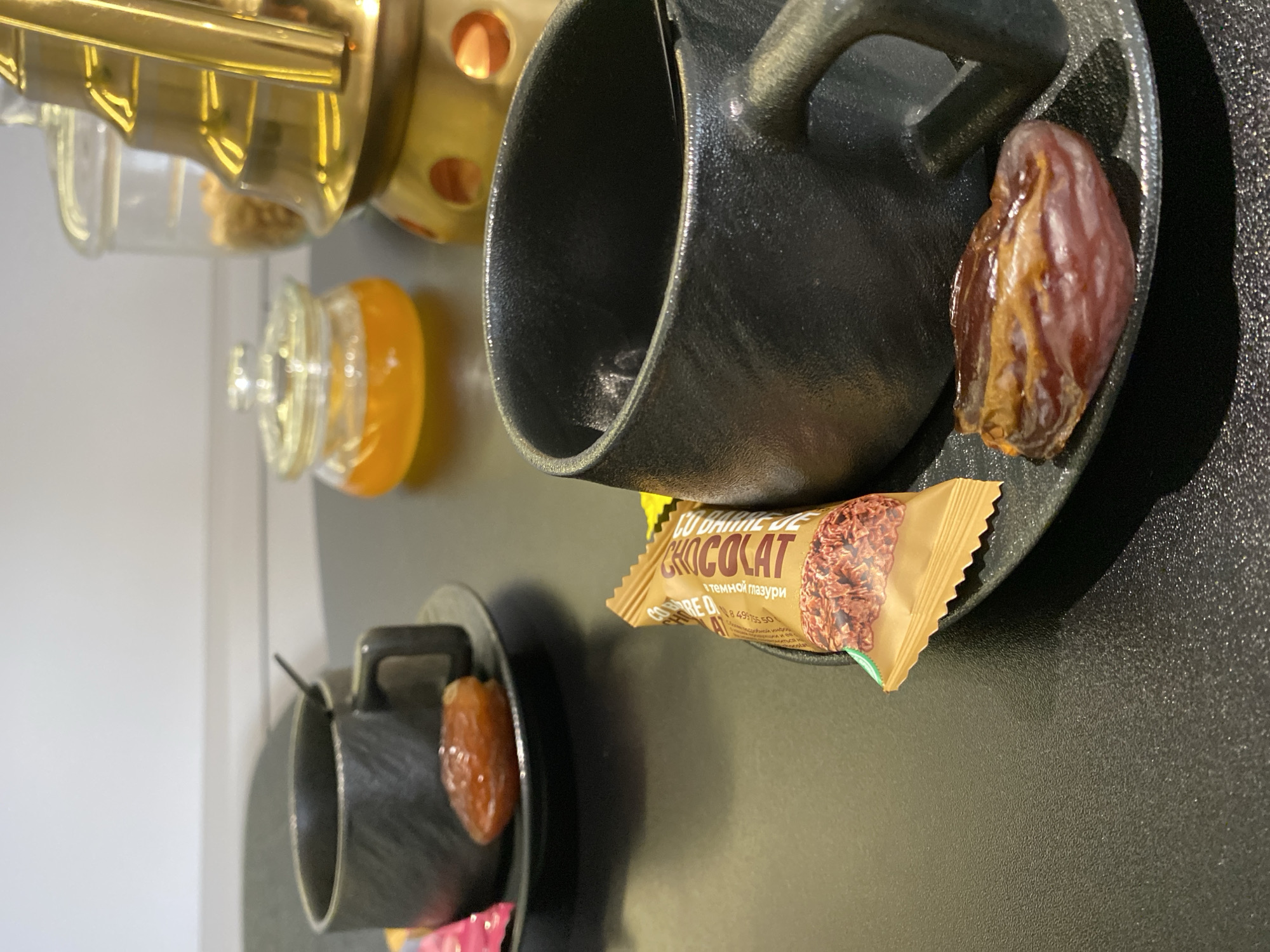 The image depicts a table setting with a mug, a date, a chocolate bar, a pot, and a jar. The mug is black and sits on a black saucer with a date resting next to it. The chocolate bar is wrapped in a tan and yellow wrapper with the word "CHOCOLAT" printed on it. A jar with a light-colored liquid inside is also visible. The pot is metallic gold in color.
The chocolate bar has "CU BAIILE DE" and "CHOCOLAT" printed on it. Underneath "CHOCOLAT" is "с темной мазури". The barcode number is 8 4917550. The image seems to be from Russia, but there is no clear information about the location in Votkinsk.
