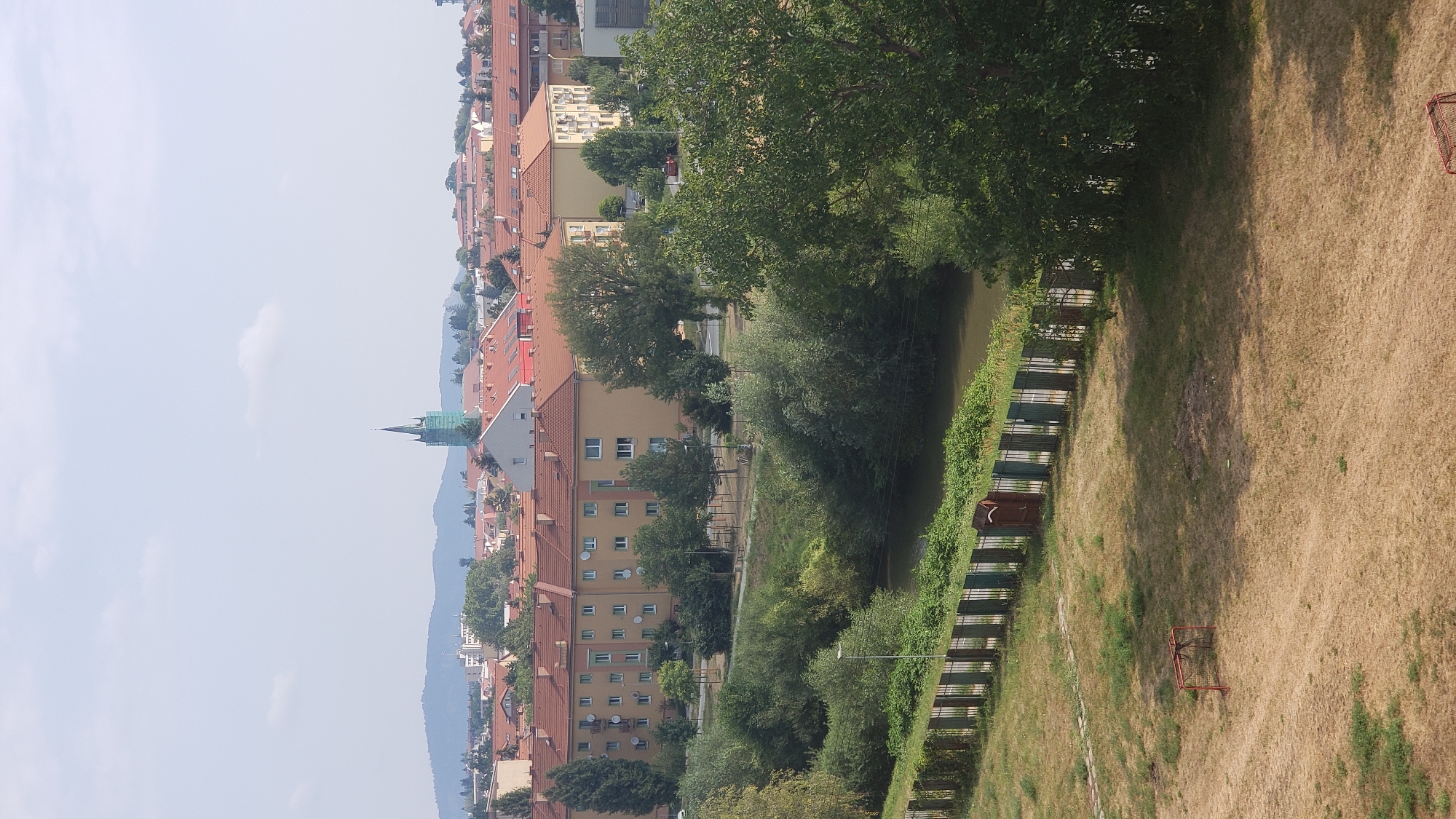 The image shows a view of the city of Prešov, Slovakia, looking towards the north. In the foreground, a dry, mowed lawn with a small, red soccer goal is visible. A metal fence separates the lawn from a body of water, likely the Sekčov river, lined with lush green trees and bushes. Beyond the trees, a row of apartment buildings with orange tiled roofs stretches across the mid-ground. Further in the distance, the cityscape includes more buildings and trees, and on the horizon, rolling hills are visible under a hazy sky with scattered clouds. A tall, green church spire with scaffolding around it stands out in the center of the distant skyline. The overall scene depicts a residential area on a clear day, with a mix of urban development and natural elements.