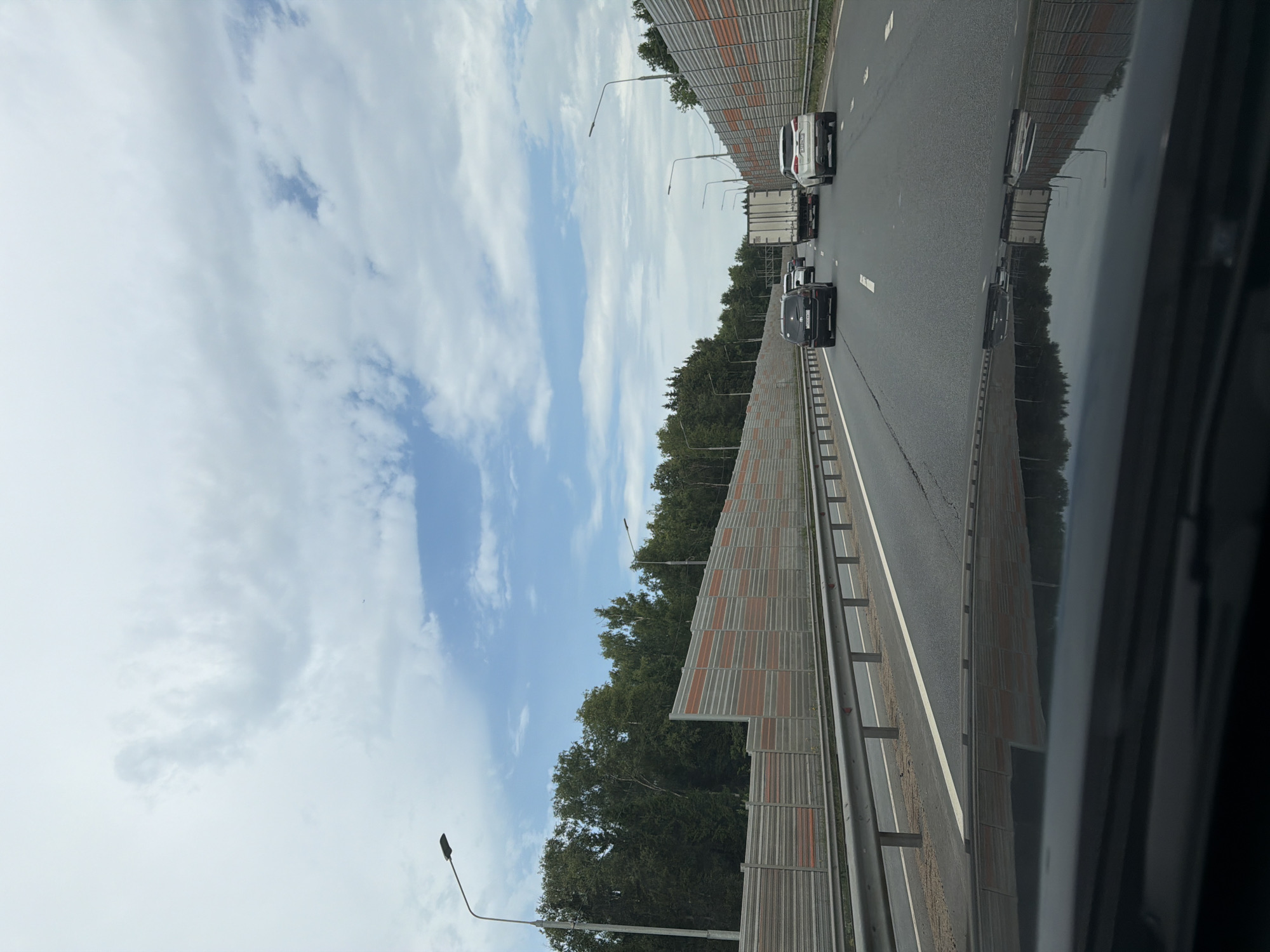 The image shows a view from inside a vehicle traveling on a highway. The highway has multiple lanes and is bordered by sound barriers on both sides, which are composed of metal panels with orange and grey sections. Beyond the sound barriers, there is a dense line of green trees. The sky is overcast with scattered clouds and patches of blue. Several vehicles are visible on the highway, including cars and a truck. The front dashboard of the vehicle taking the photo is reflected in the lower part of the image. The scene appears to be a typical urban or suburban highway environment. No specific landmarks indicating the city of Mar'ino, Russia are visible.