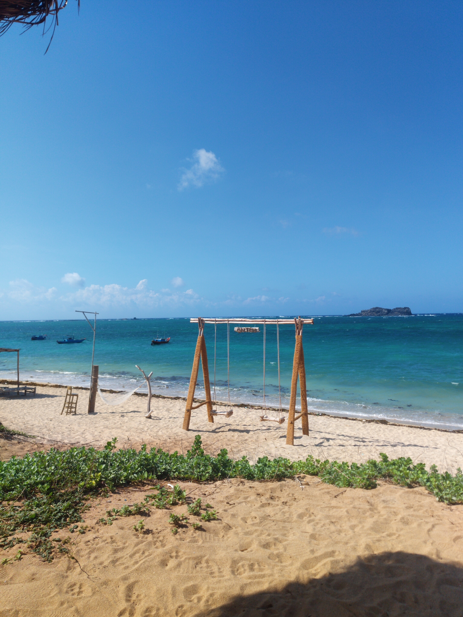 The image depicts a tranquil beach scene in Phú Quý Special Administrative Region, Vietnam. The scene is dominated by a clear blue sky and turquoise water. In the foreground, a sandy beach is covered with vegetation. A wooden swing set with rope swings stands on the beach. In the water, a small rocky island is visible. Several boats are at anchor in the distance. A hammock is seen between two poles.