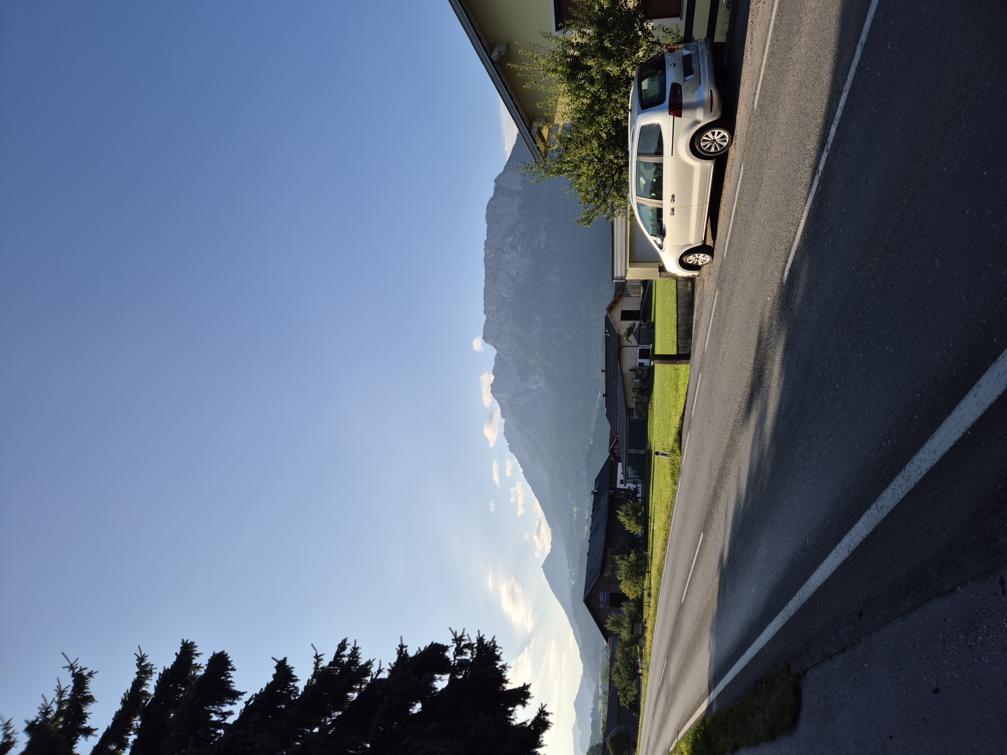 The image depicts a daytime scene in Niederndorf, Austria, featuring a road, a silver minivan, houses, green fields, and a mountain range in the background. A large conifer tree is visible in the foreground on the left. The road is paved and has a white line marking its edge. The minivan is parked on the right side of the road. Houses with yellow and brown facades are situated behind the minivan and across the road. The mountains in the distance are covered in vegetation and appear to be illuminated by the sun. The sky is clear and blue, with a few wispy clouds. The overall impression is one of a rural or semi-rural setting on a sunny day.