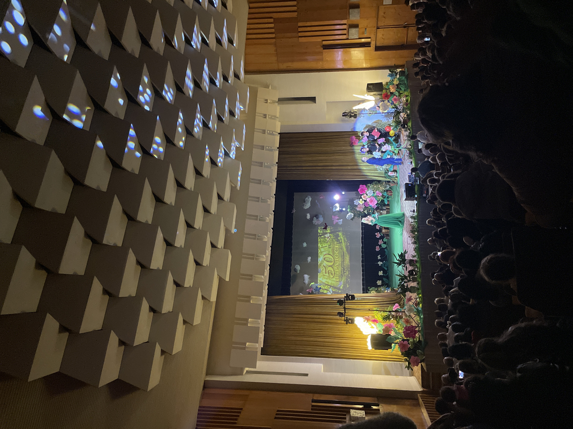 The image depicts an indoor event taking place in an auditorium or theater, likely in Zlatograd, Bulgaria. The scene is dominated by a brightly lit stage on the right, contrasted with a dimly lit audience area and a geometrically patterned wall on the left.
The stage is adorned with lush floral decorations featuring numerous artificial flowers in shades of pink, purple, white, and yellow, alongside green foliage. A large screen at the rear of the stage displays a visual, possibly a forest or nature scene with butterflies or moths. Visible text on the screen includes "50 МИ" and "РАДОСТ," likely part of the phrase "50 МИГОВ РАДОСТ" (50 Moments of Joy). One individual, dressed in a long, flowing green gown, stands on the stage, facing the audience, suggesting they are a performer or presenter.
The audience, positioned on the far right, is largely obscured by darkness, with only the tops of their heads visible. Many attendees are seen holding up cell phones, indicative of them recording or photographing the event.
To the left, a prominent architectural feature is a wall covered with numerous light-colored, three-dimensional geometric shapes, possibly acoustic panels or decorative elements. These shapes are illuminated by projected lights, creating abstract patterns of blue and white dots and streaks that add to the visual ambiance.
The setting suggests an evening event, given the reliance on artificial lighting to illuminate the stage and architectural features. The overall atmosphere points to a cultural performance, celebration, or public address.