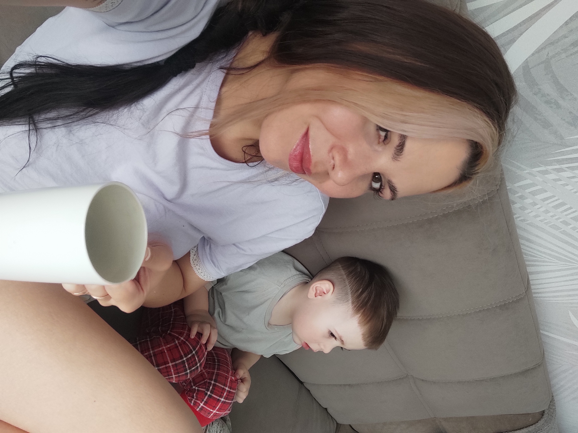 The image shows a woman and a young child on a gray upholstered couch. The woman has dark brown hair with a blonde streak in the front and is wearing a light purple t-shirt with lace detailing at the sleeves. She is holding a white cylindrical object, possibly a mug, in her hand. She is looking at the camera. The child has a brown bowl cut haircut and is wearing a gray t-shirt and red plaid pants. The child is looking down and to the right. The background includes a patterned wall. The location is Chaikovsky, Russia.
