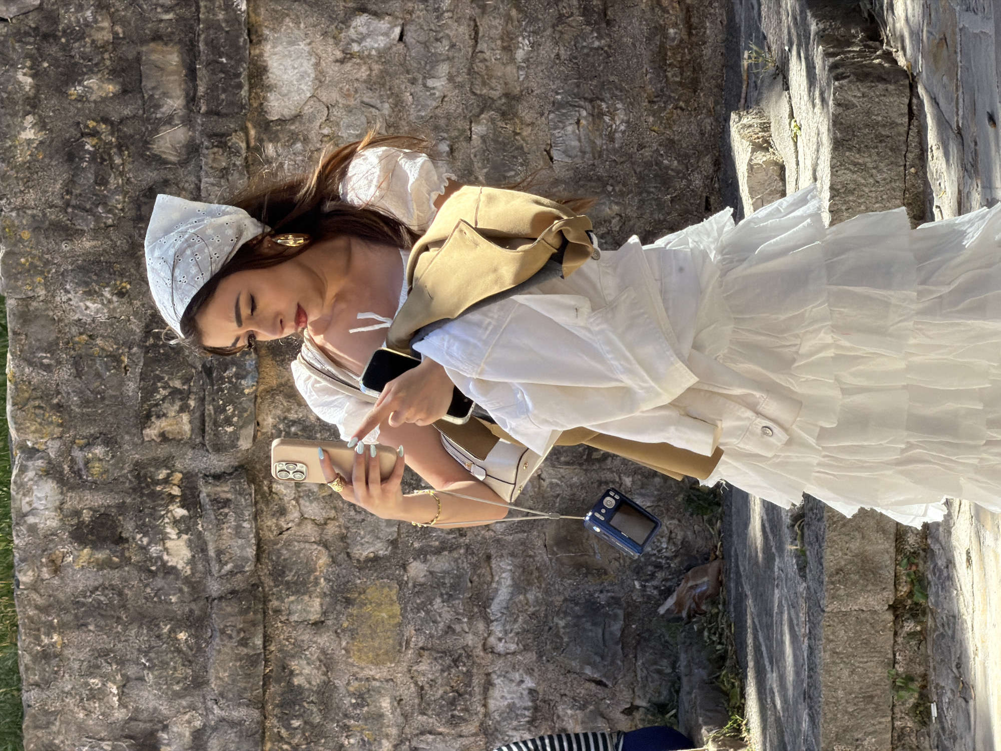 A woman is standing on a stone step in Budva, Montenegro. She is wearing a white dress, a tan jacket, and a white headscarf. She is holding a gold smartphone in her left hand, pointing at the screen with her right index finger. In her right hand, she holds a small blue digital camera connected to the phone by a strap. Her hair is brown. She wears gold earrings and rings. The background is a stone wall. Light is to the right of the photo and casts a shadow on the scene.