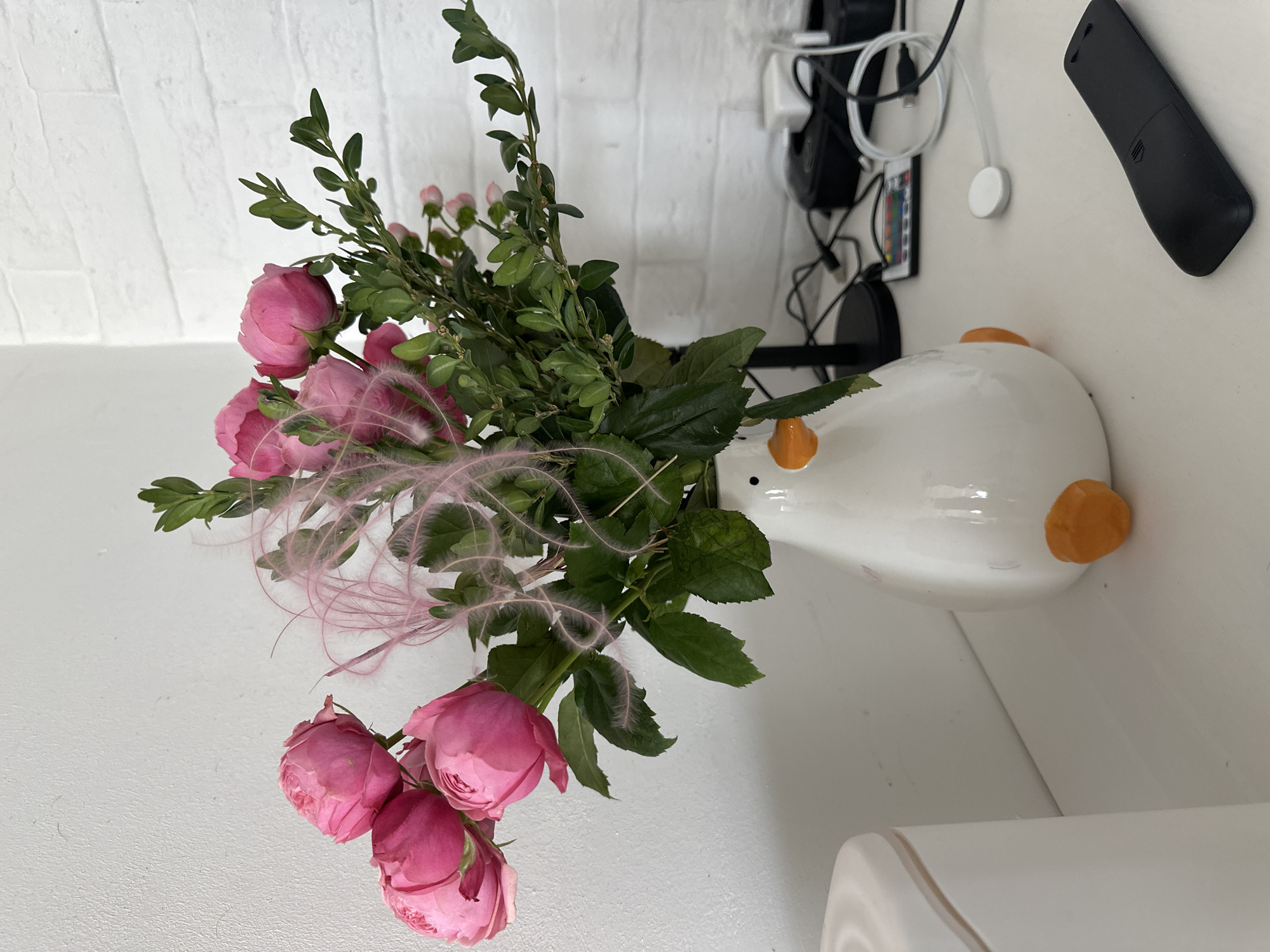 A bouquet of pink roses and greenery is arranged in a white duck-shaped vase on a white surface. The setting appears to be indoors. There is a white brick wall in the background, along with electronic devices and cables mounted on the wall. Moscow, Russia is not apparent in the image.