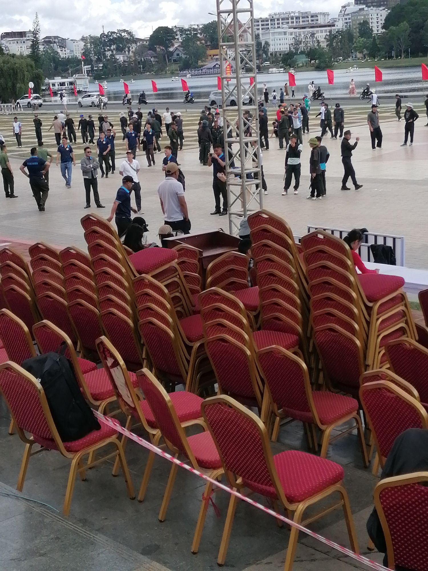 The image depicts an outdoor event taking place in Da Lat, Vietnam, likely at a public square or park adjacent to Xuan Huong Lake. In the foreground, numerous red upholstered chairs with gold metal frames are stacked or arranged in rows. Many chairs are empty, while some have a black backpack on them. Red and white striped caution tape is strung between some of the chairs.
In the mid-ground, a large gathering of people are present. Many individuals are dressed in dark uniforms, suggesting they may be security personnel or participants in an official event. Some people are standing, while others appear to be walking or conversing. A few individuals are dressed in casual civilian clothing.
The background reveals a city landscape with buildings, trees, and a body of water, consistent with the scenery around Xuan Huong Lake. Red flags are visible, planted along the waterfront and on structures. A large metal scaffolding structure is present on the left side of the image, indicating a stage or platform for the event. The sky is overcast.