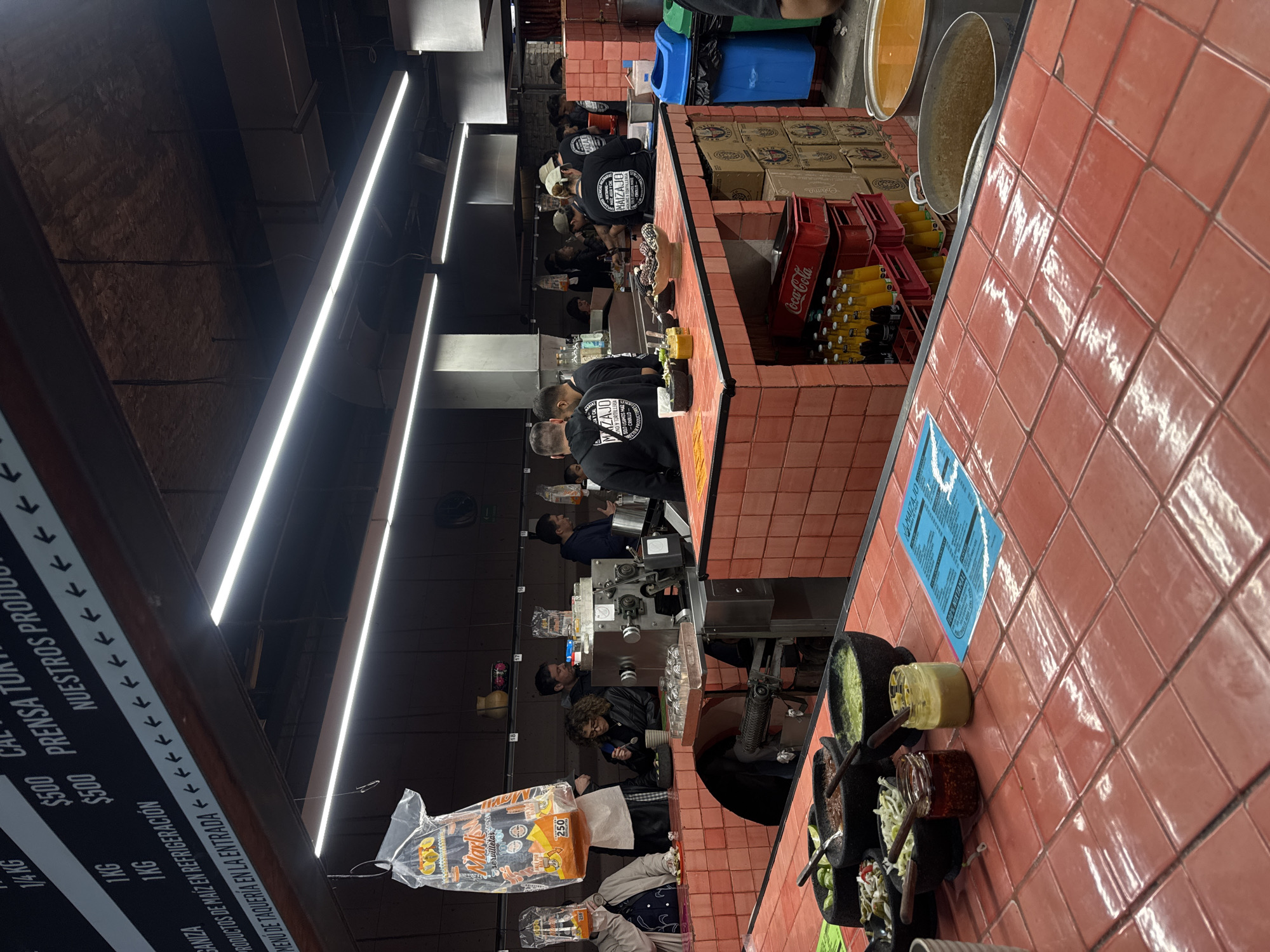 The image shows the interior of a Mazajo taqueria in Mexico City, Mexico. A long, tiled counter stretches into the distance, where employees in black shirts with the Mazajo logo are preparing food. Coca-Cola bottles in red crates are stacked near the counter. Patrons stand in front of the counter to order. Bowls of salsa and condiments line the edge of the counter closest to the camera. Packaging of tortillas with "Marl Servilletas" visible. A sign on the left lists prices for various products including "Ganiza" and "Prensa Tort". A series of fluorescent lights illuminate the scene.