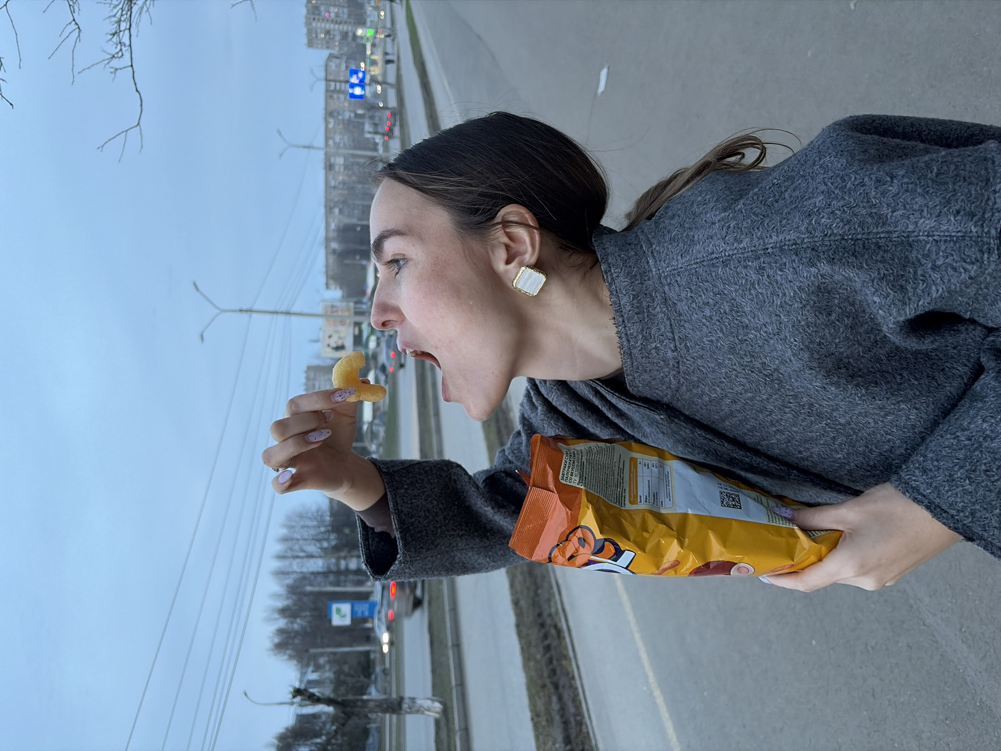 A woman with dark hair pulled back is holding a bag of snacks and a single snack in her hand, poised to eat it. She is wearing a gray coat and has a square earring. She is standing on a sidewalk next to a road. In the background are buildings and street lights, indicating an urban environment. A road sign, visible in the background, indicates this location is in Izhevsk, Russia.