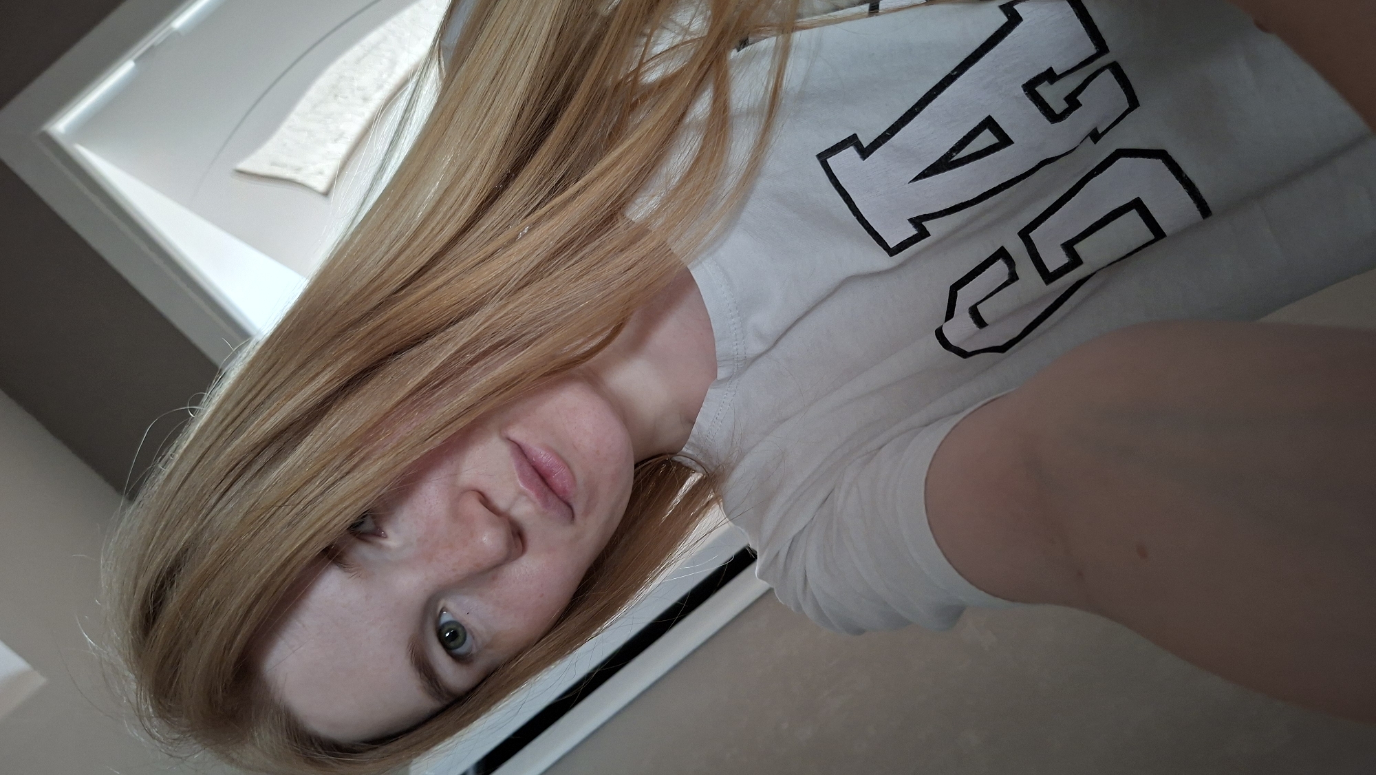 A young woman with long, blonde-red hair and fair skin with freckles is captured in a close-up, selfie-style shot. She is wearing a white t-shirt with the black letters "GAP" printed across it. Her facial expression is relaxed, with her eyes looking towards the camera and her lips slightly parted. The background appears to be an indoor setting, possibly a room in Chaikovsky, Russia, with a doorway and a wall visible. The lighting suggests it is daytime, possibly near a window.