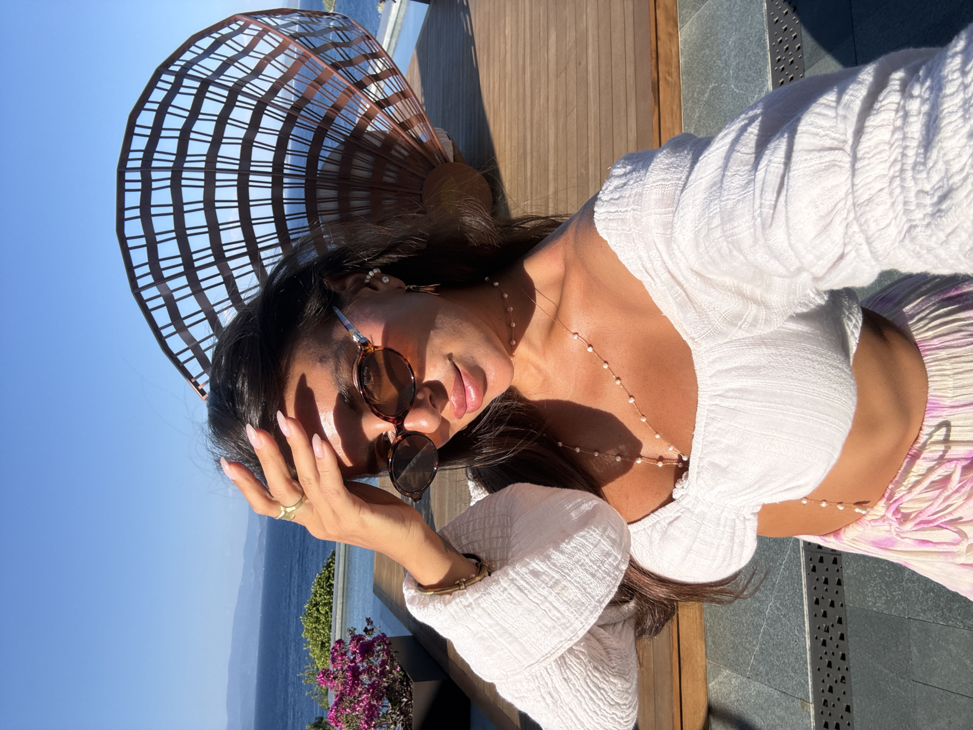 A woman is taking a selfie outdoors in Bodrum, Turkey. She is wearing sunglasses and a white top with flowing sleeves, and a pearl necklace. Her hand is on her forehead, shielding her eyes from the sun. In the background, a large, fan-shaped sculpture stands near a body of water, likely the Aegean Sea, with blue skies overhead.