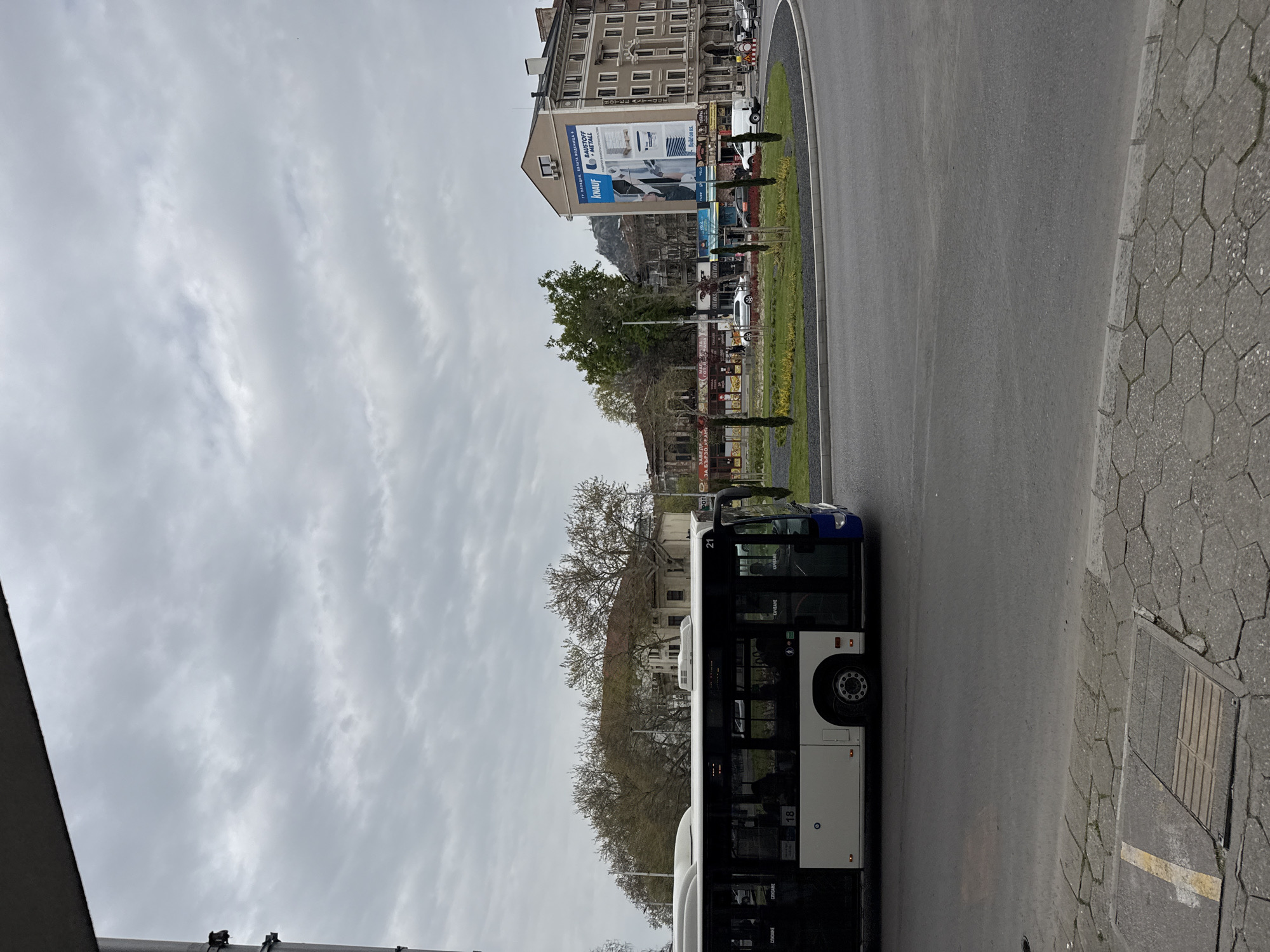 The image shows a city street in Plovdiv, Bulgaria, with a white and blue bus on the right side of the road. A sidewalk made of hexagonal pavement is also on the right. Across the street, there is a circular grassy area and a multi-story building with a large advertisement on its side. The sky is overcast. No people are visible in the image.