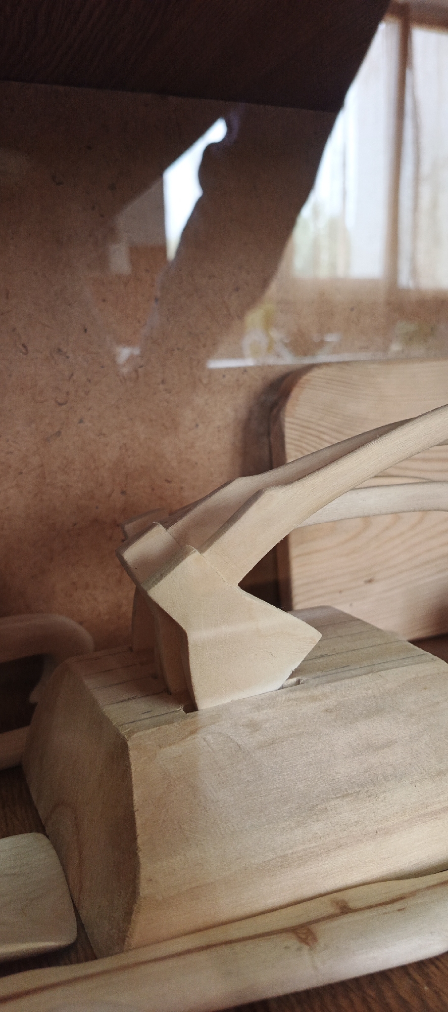 The image depicts a wooden axe resting in a wooden block. It is located in Кемуль, Russia. The axe head is triangular, and the handle is long and slender. The block is a rough cube shape. The wood is light-colored and appears unfinished. There are other pieces of wood in the background. The setting seems to be an indoor workshop or craft area.