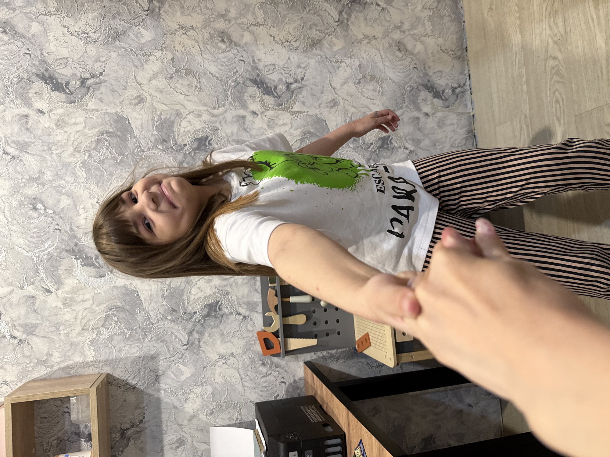 An indoor, vertically oriented shot depicts a young girl extending her hand towards the camera, her hand meeting another person's hand in the foreground. The girl, with light brown hair and bangs, is smiling and looking at the camera. She wears a white t-shirt with a bright green, paint-splatter-like design and black text that appears to read "ESCAPADE" and "PARIS" (with stylized letters, some resembling Cyrillic characters like 'Д', 'А', 'Р', 'И', 'Ж'). She is also wearing black and beige striped pants.
The background features a textured wall with a gray and white, marble-like or distressed plaster pattern. To the right, there is light wood-patterned flooring. On the left side of the room, a light wooden cubby shelf is visible. On the right, a dark-colored desk or table holds a black printer (identifiable by the "EPSON" logo). Above the printer and behind the girl, a gray pegboard with several wooden toy tools (including a saw, wrench, and roller) is mounted on the wall, suggesting a play area. Another wooden toy or device is on the table next to the pegboard. The scene is brightly lit, likely by indoor lighting, and conveys a cheerful and interactive moment, possibly of holding hands. The location is a room in a home in Rusanovo, Russia.