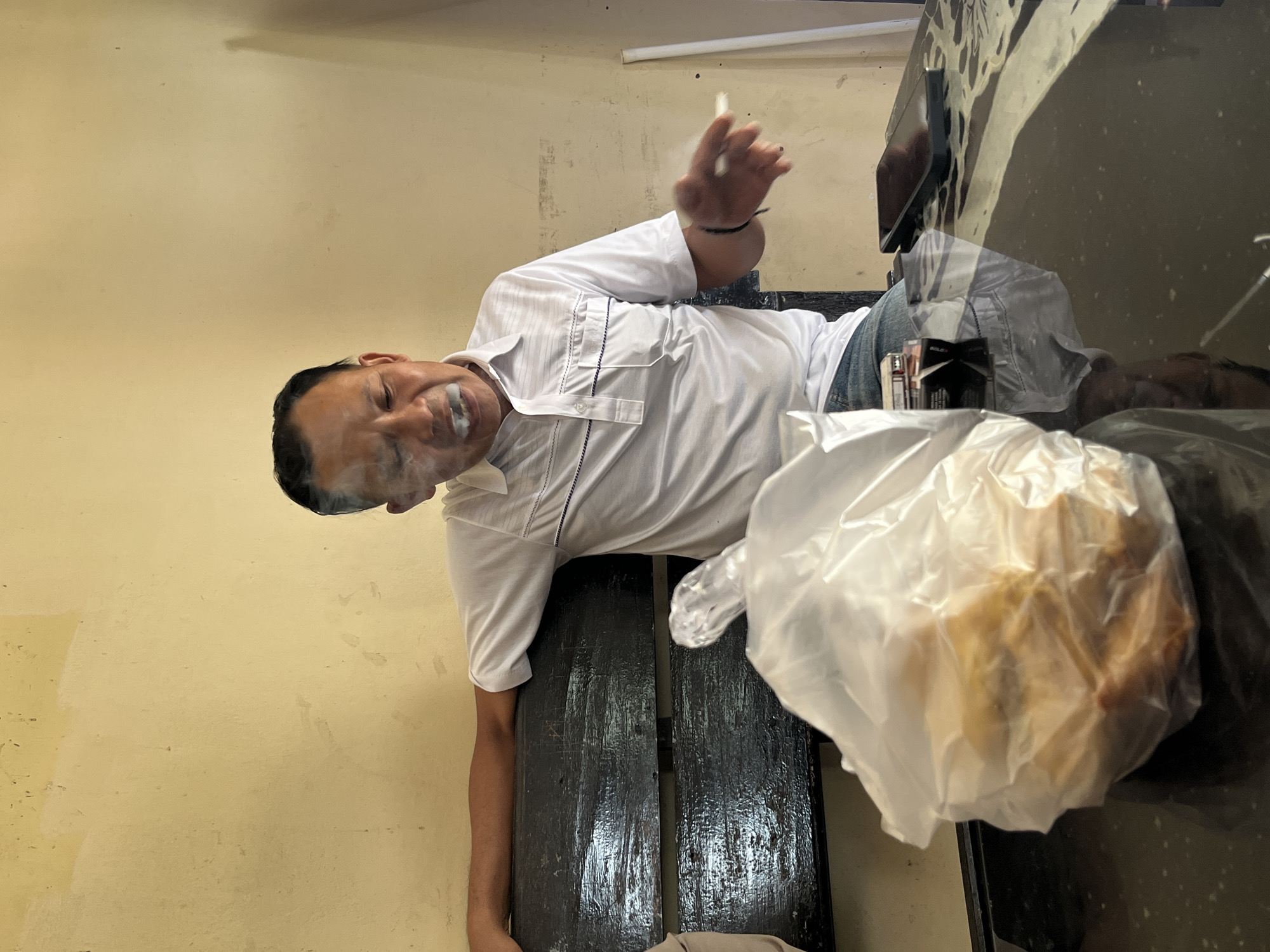 In Tanakawu, Indonesia, a man in a white polo shirt is seated on a dark wooden bench, exhaling smoke from a cigarette. A plastic bag containing food items is placed on a black, reflective table in front of him. A smartphone and a pack of cigarettes are also visible on the table. The background consists of a yellow-painted wall with visible scuff marks and a white vertical object. The lighting suggests an indoor environment with natural light.