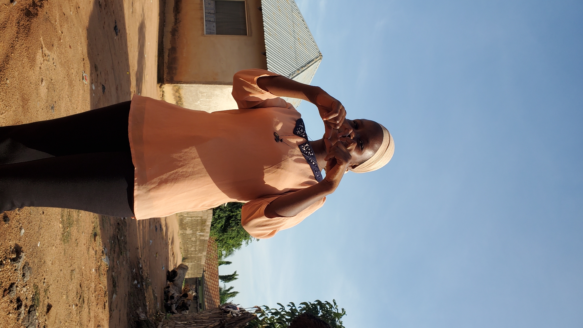 A woman in a light peach blouse and dark leggings stands outside a modest building with a corrugated metal roof. She wears a light beige headscarf and forms a heart shape with her hands in front of her face. The ground is dusty and sandy, with scattered debris. In the background, other structures and trees are visible under a clear blue sky. The scene appears to be in a residential area within the city of Jalingo, Nigeria.