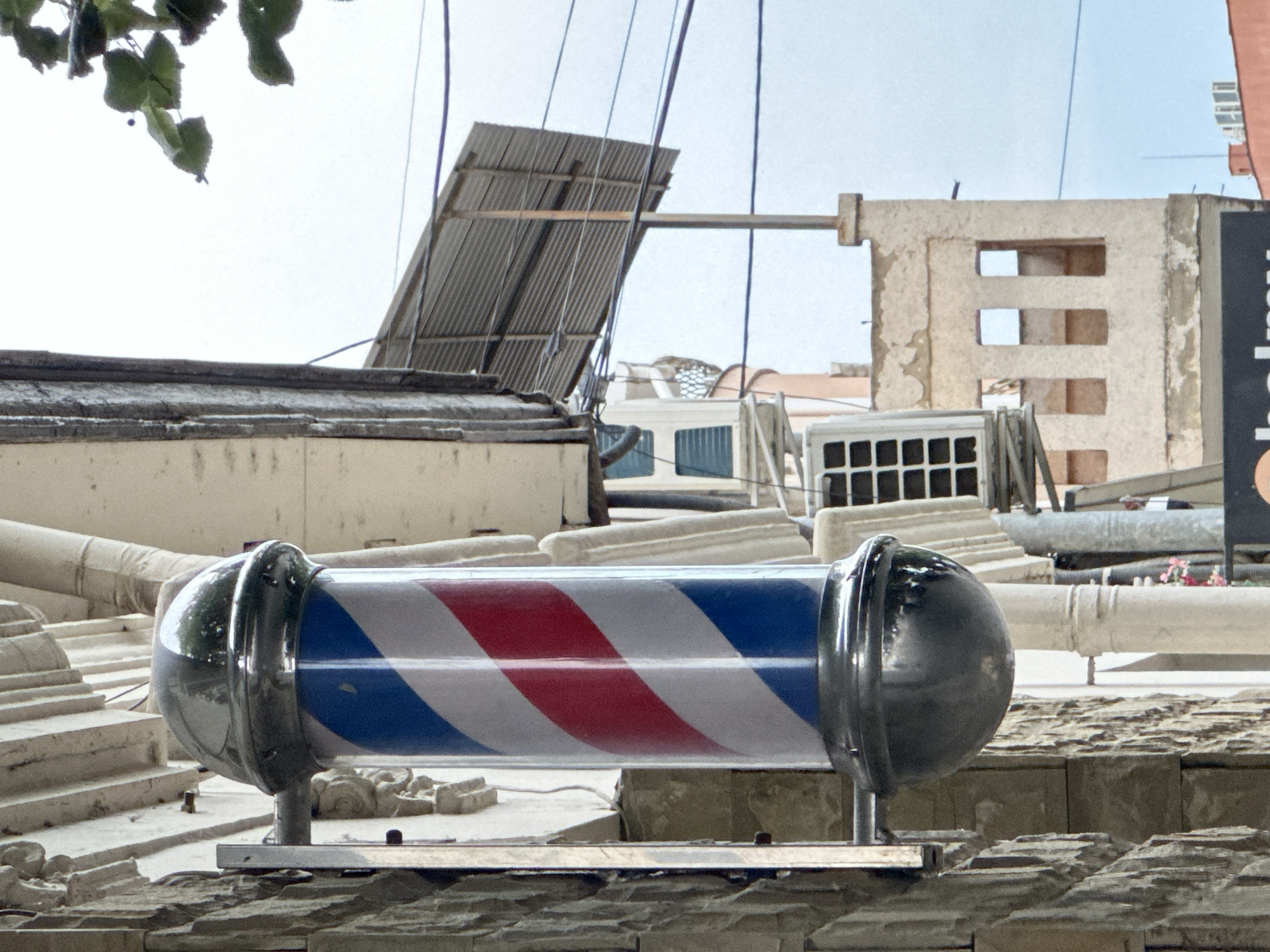 The image shows a barber pole mounted on a stone wall building. The barber pole is a traditional red, white, and blue striped cylinder with a chrome top and bottom. In the background, there is a corrugated metal roof extending from another building, power lines, an air conditioning unit, and a building with square window openings. A few green leaves from a tree are visible in the upper right corner. The scene appears to be on a street in a city. The specific location of the barber pole on Korzo Street in Uzhhorod, Ukraine, is identified based on contextual information not directly discernible from the image alone.