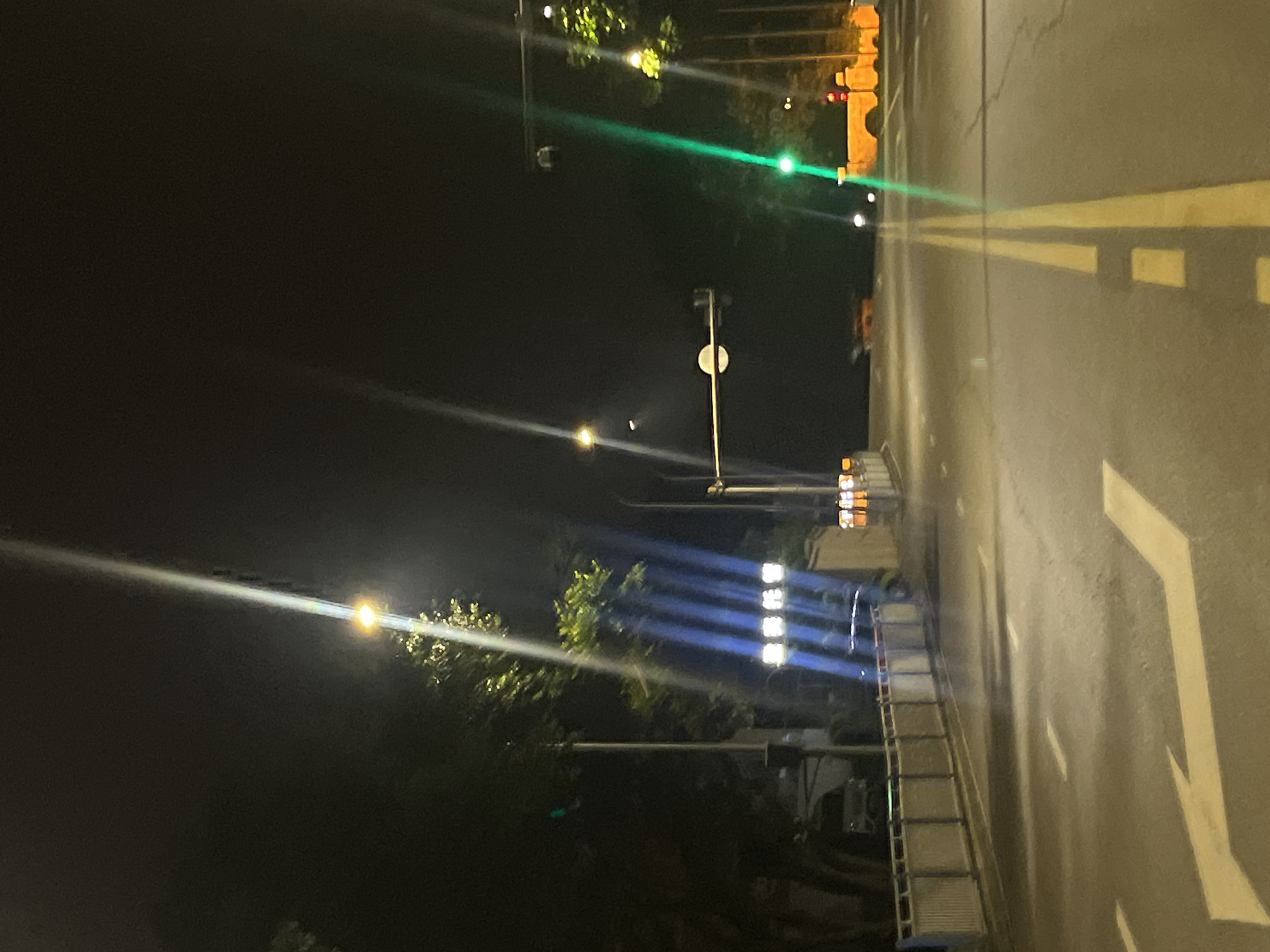 The image depicts a night scene on a street in Jiulongpo District, China. The road, paved with asphalt, is marked with yellow and white lane dividers and directional arrows. Streetlights illuminate the scene, casting long beams of light across the road. On the left side of the road, a white metal barrier runs along the sidewalk, backed by trees and a few vehicles. In the distance, a traffic light with a green light is visible, and to the right, a structure with illuminated yellow lights is present. A security camera is mounted on a pole above the road. The sky is dark, indicating nighttime. There are no people clearly visible in the frame.