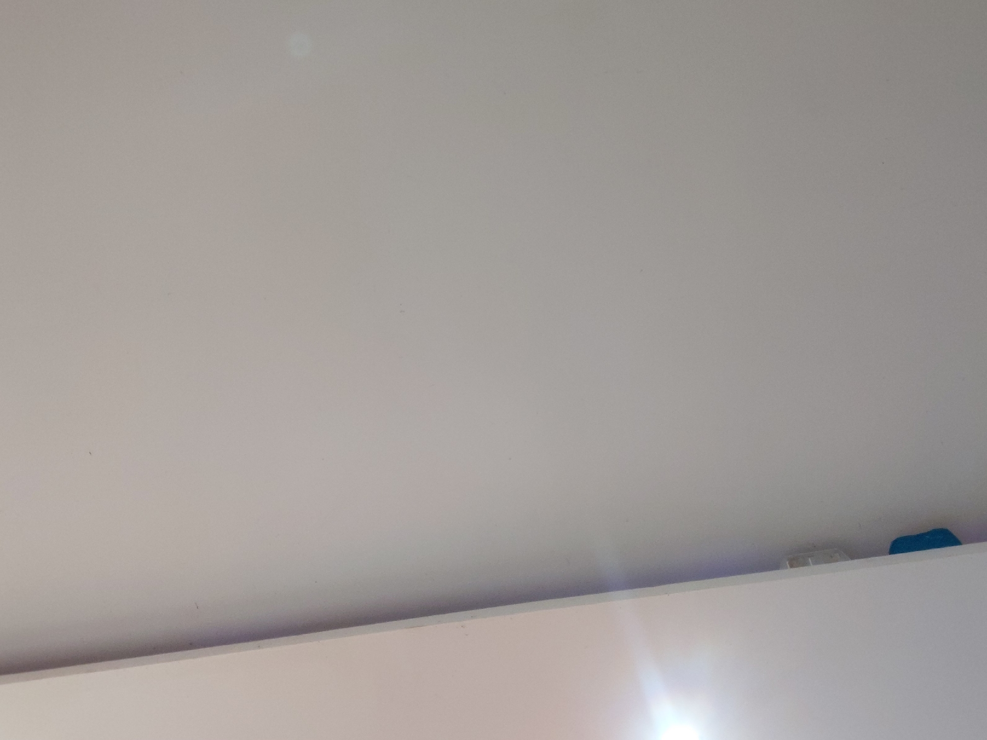 The image captures an interior scene, primarily depicting a light-colored ceiling or upper wall, which appears to be off-white or light gray. No individuals are visible. The majority of the frame is dominated by this plain, smooth surface, showing only minor specks or blemishes upon close inspection.
A distinct horizontal line runs across the bottom third of the image, marking the division between the upper surface and a lower, also light-colored surface, possibly the top of a wall, a shelf, or a piece of furniture. In the bottom right corner, resting on this lower surface, are two small items: one is a light-colored, possibly rectangular object, and beside it, a brightly colored, irregularly shaped blue object, possibly plastic.
A strong light source, creating a prominent lens flare, is visible in the bottom right of the image, intensely illuminating the lower surface. This suggests strong artificial lighting or direct sunlight entering the space. The overall ambiance indicates an indoor setting. There is no visible text present in the image, and no specific visual cues within the image itself confirm the location as Santa Ana, Venezuela.