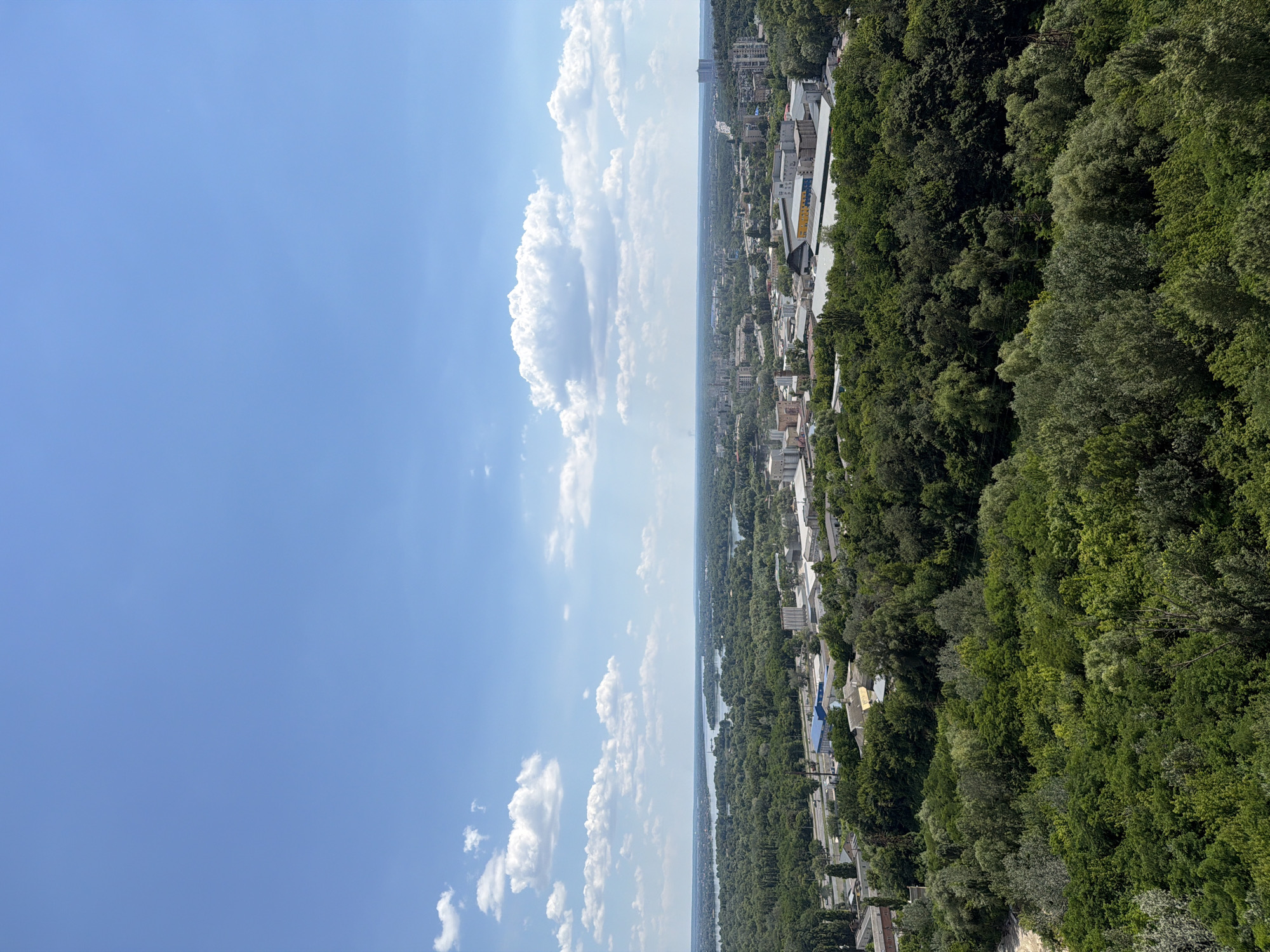 This media file depicts a panoramic view of a city, likely Kyiv, Ukraine, on a clear, sunny day. The image is oriented vertically, presenting a vast expanse of blue sky with scattered fluffy white clouds on the left side, taking up about half of the frame. On the right side, a densely forested hillside slopes down towards a city and a body of water, likely a river.
The city appears to be built on a plain stretching along the riverbank. Various buildings, ranging from smaller structures with light-colored roofs to larger, more modern multi-story buildings, are visible. Some structures have distinctive yellow or blue accents. The dense green foliage of the forest covers the hillside, creating a stark contrast with the urban landscape and the bright sky. The river meanders through the landscape, reflecting the sky and the surrounding greenery.
The overall impression is one of a vibrant city integrated with its natural surroundings. The time of day appears to be late morning or early afternoon, judging by the bright sunlight and the clear sky. There are no visible people or specific objects of focus within the scene, suggesting it's a landscape shot. No text is discernible in the image. The bright, clear weather and the expansive view evoke a sense of peace and natural beauty.
