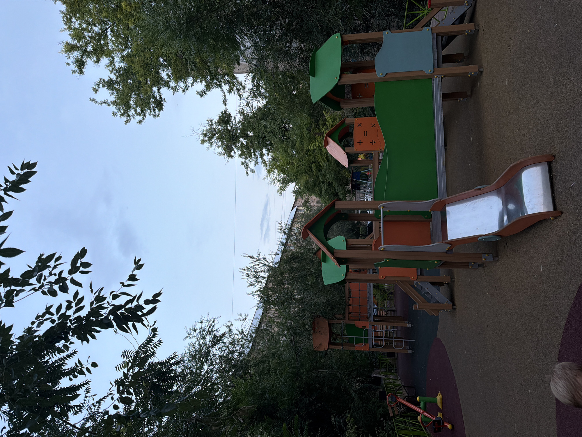 The image shows an outdoor playground scene in the city of Shymkent, Kazakhstan, on a cloudy day. In the foreground, a toddler with blonde hair is partially visible, lying on a reddish-purple soft play surface. The main focus is a multi-level wooden playground structure with green and orange accents. This structure features slides, stairs, and covered areas. To the left, a red and yellow seesaw is positioned on a maroon soft play surface. Trees and foliage surround the playground, with a multi-story building visible in the background through the trees. The sky is overcast with pale blue and gray clouds.