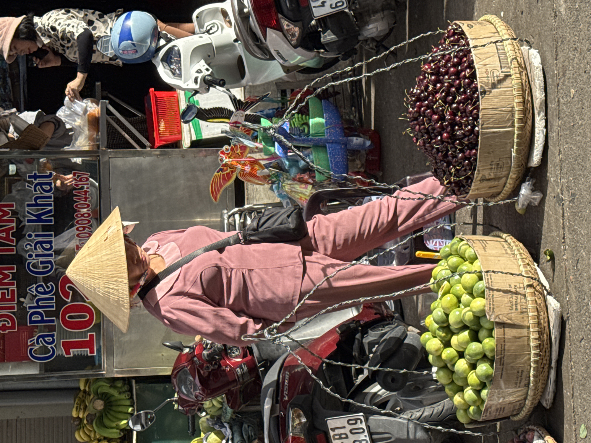 A woman wearing a conical hat and pink clothing walks through an outdoor market in Da Lat, Vietnam, carrying two baskets of fruit suspended from a pole. She is positioned on the right side of the frame, with her back slightly turned towards the viewer. The basket on her left holds a large pile of green citrus fruits, likely limes, and the basket on her right is filled with dark red cherries. The fruits are displayed in woven baskets that are supported by a metal frame. In the background, there are motorcycles parked, a market stall with signage in Vietnamese, and other market items such as bananas and colorful toys. The ground is paved, and the scene is illuminated by sunlight.